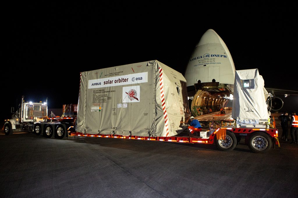 A large transport truck delivers the European Space Agency’s Solar Orbiter spacecraft in a protective container to a cargo plane at night. The aircraft, operated by Volga-Dnepr Airlines, has its nose lifted open to reveal the spacious cargo hold, which is illuminated as workers prepare for loading. The container is labeled with “Airbus,” “Solar Orbiter,” and “ESA” logos, indicating the mission’s collaboration.