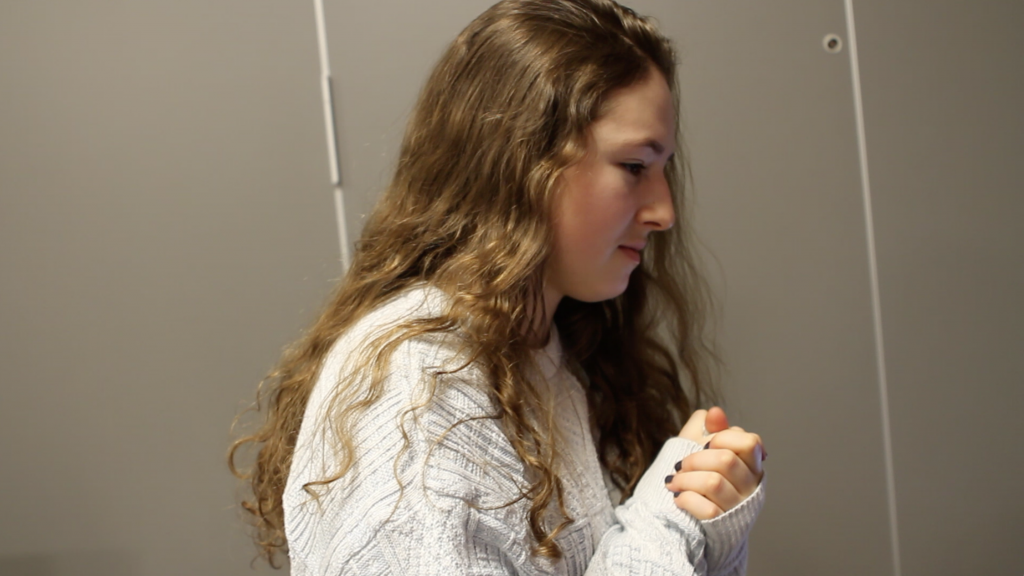A young woman with long, wavy brown hair, wearing a light knit sweater, is sitting indoors with her hands clasped together, looking intently to the side. The background is plain and gray.
