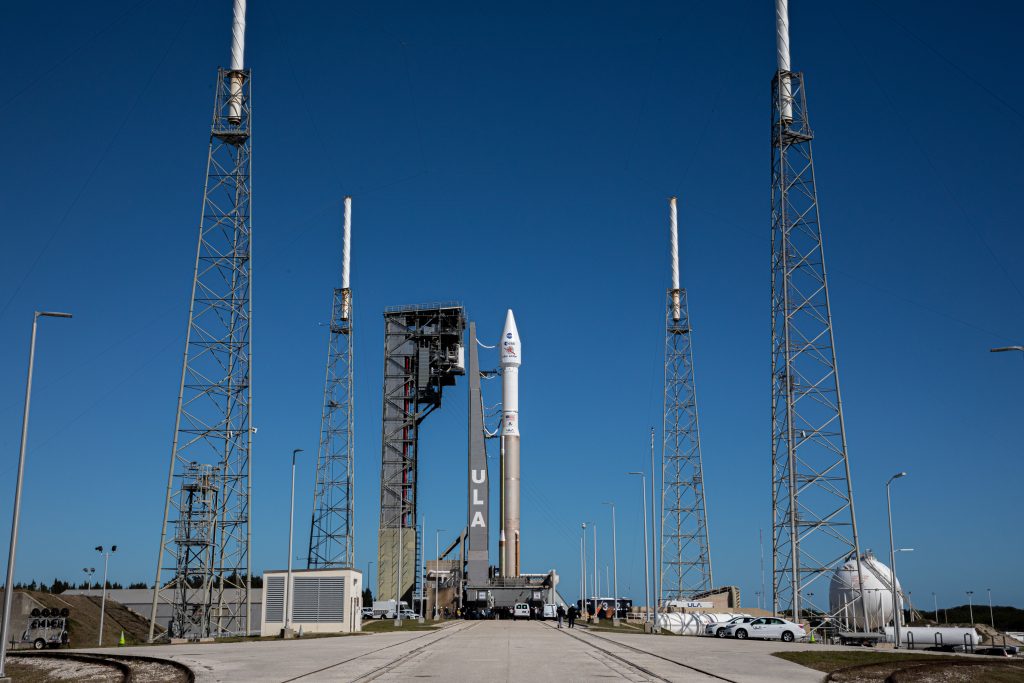 A United Launch Alliance (ULA) Atlas V rocket stands vertically on the launch pad under a clear blue sky. The rocket is flanked by four tall lightning towers and connected to a gray service structure. Ground vehicles and support equipment surround the base, and NASA and ESA logos are visible on the rocket's payload fairing.