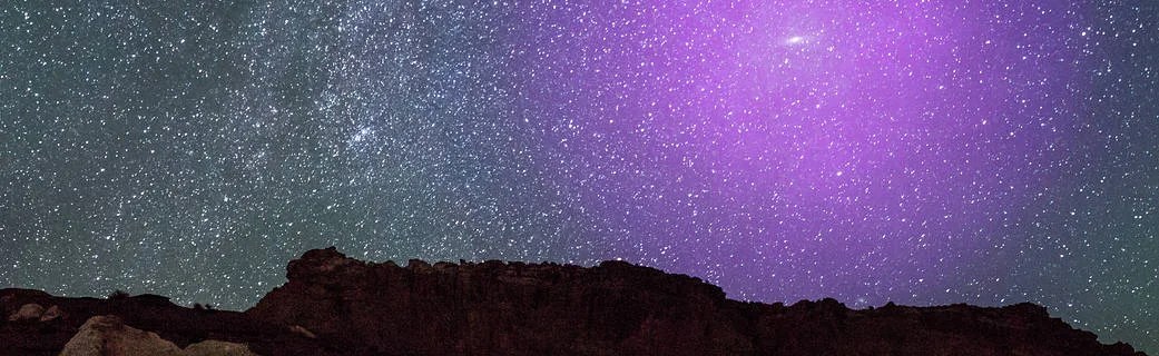 Bottom half of image: black silhouette of the landscape. Above the silhouette is a star-filled sky against a bluish-grey background. Right half of the sky is glowing purple-pink.