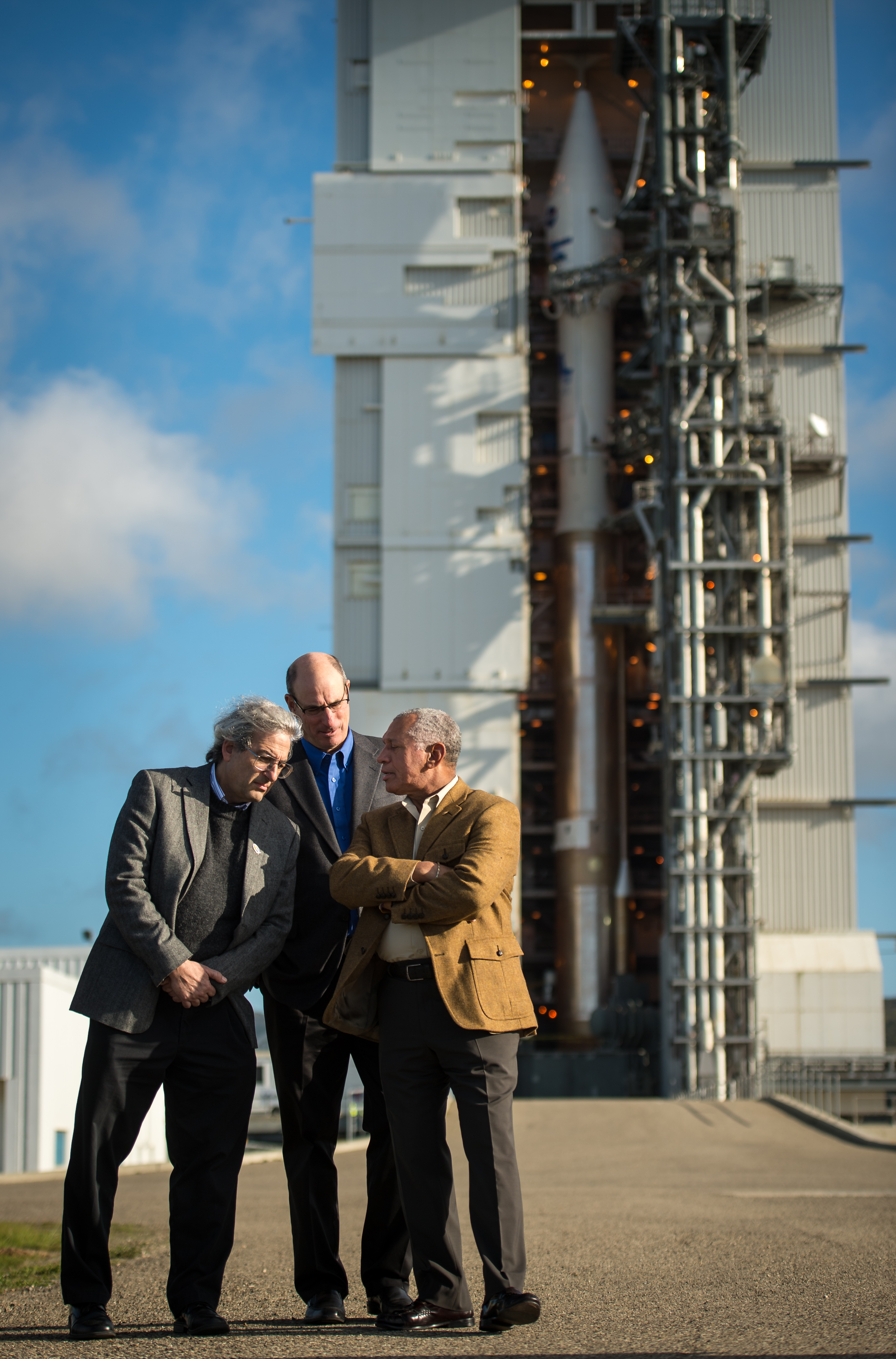 Three men are in close conversation in front of a rocket standing vertical in a hangar.
