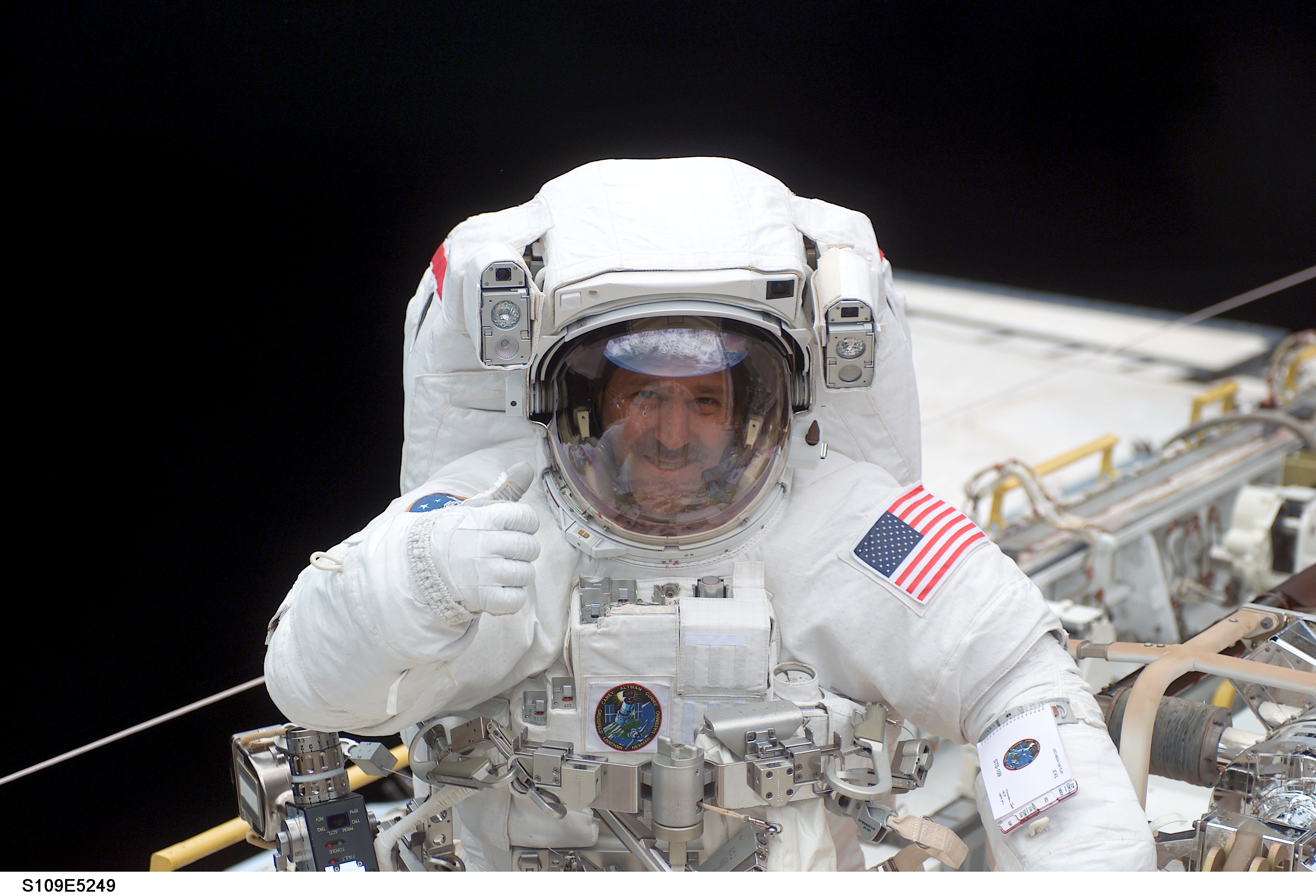 John Grunsfeld stands in his spacesuit during a Space Shuttle mission. He is giving the thumbs up and smiling for the camera.