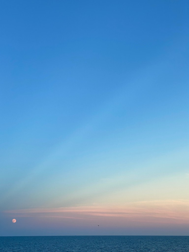 The Moon hangs low over the ocean against a blue and pink sunset sky