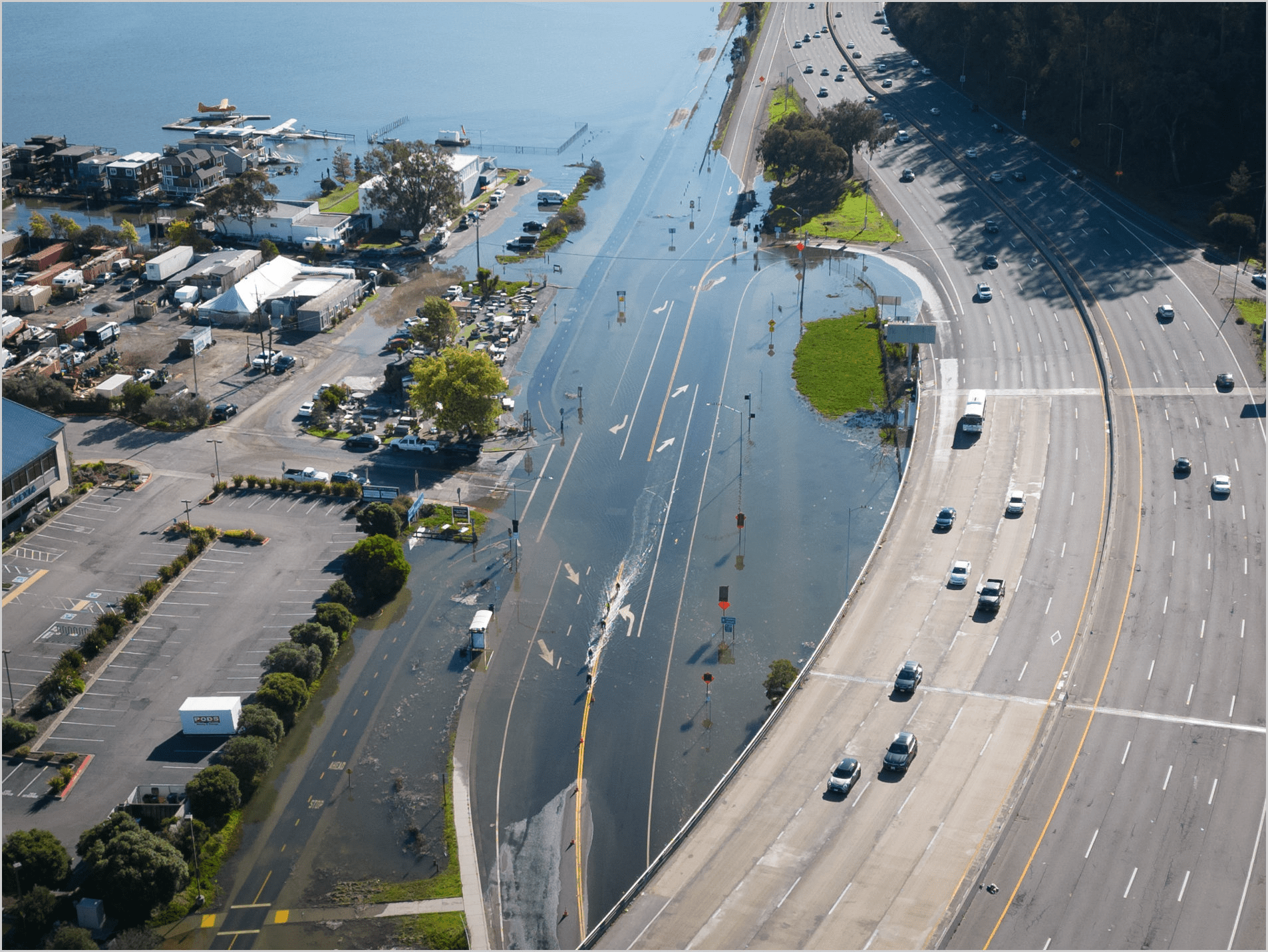 flooded roadway by large coastal highway onramp (right) and various small buildings and parking on the coast side (left)