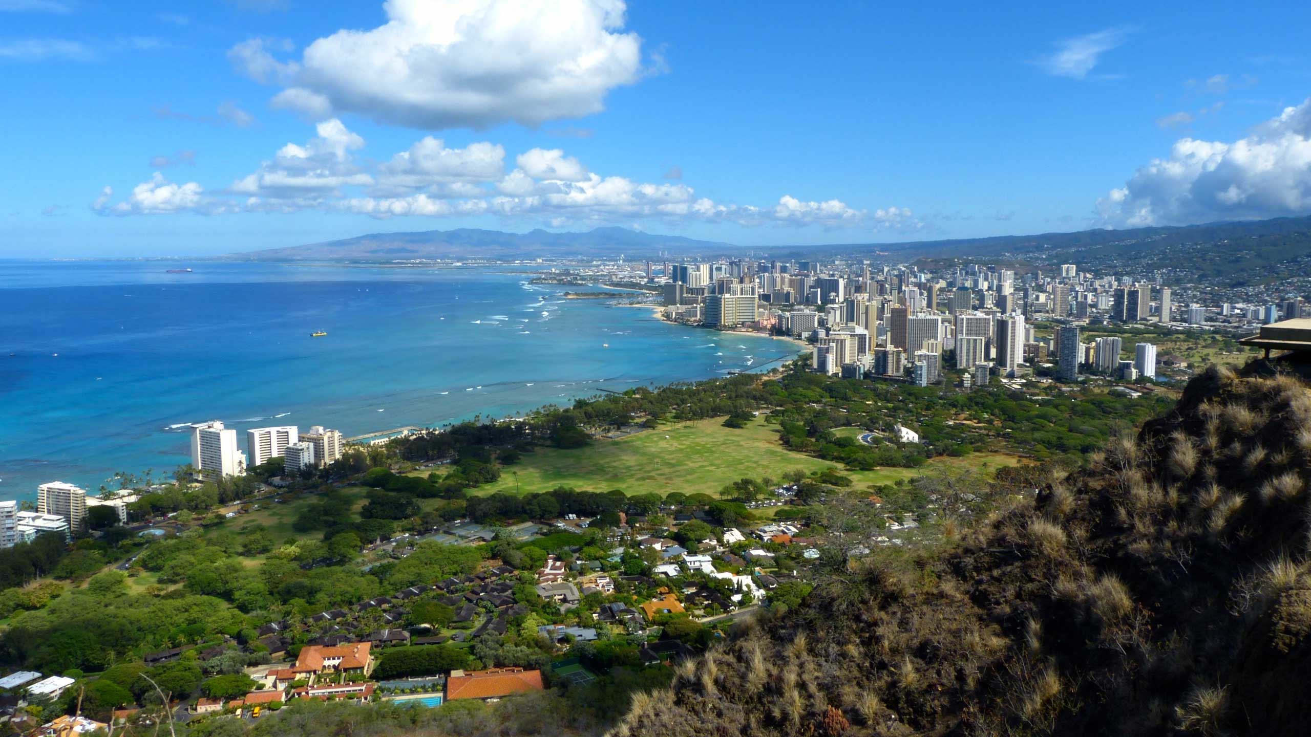 Photo of the city of Honolulu, from a nearby peak. Skyscrapers are featured right, with sprawling fields in the foreground with the ocean expanse on the left under a mostly sunny sky.
