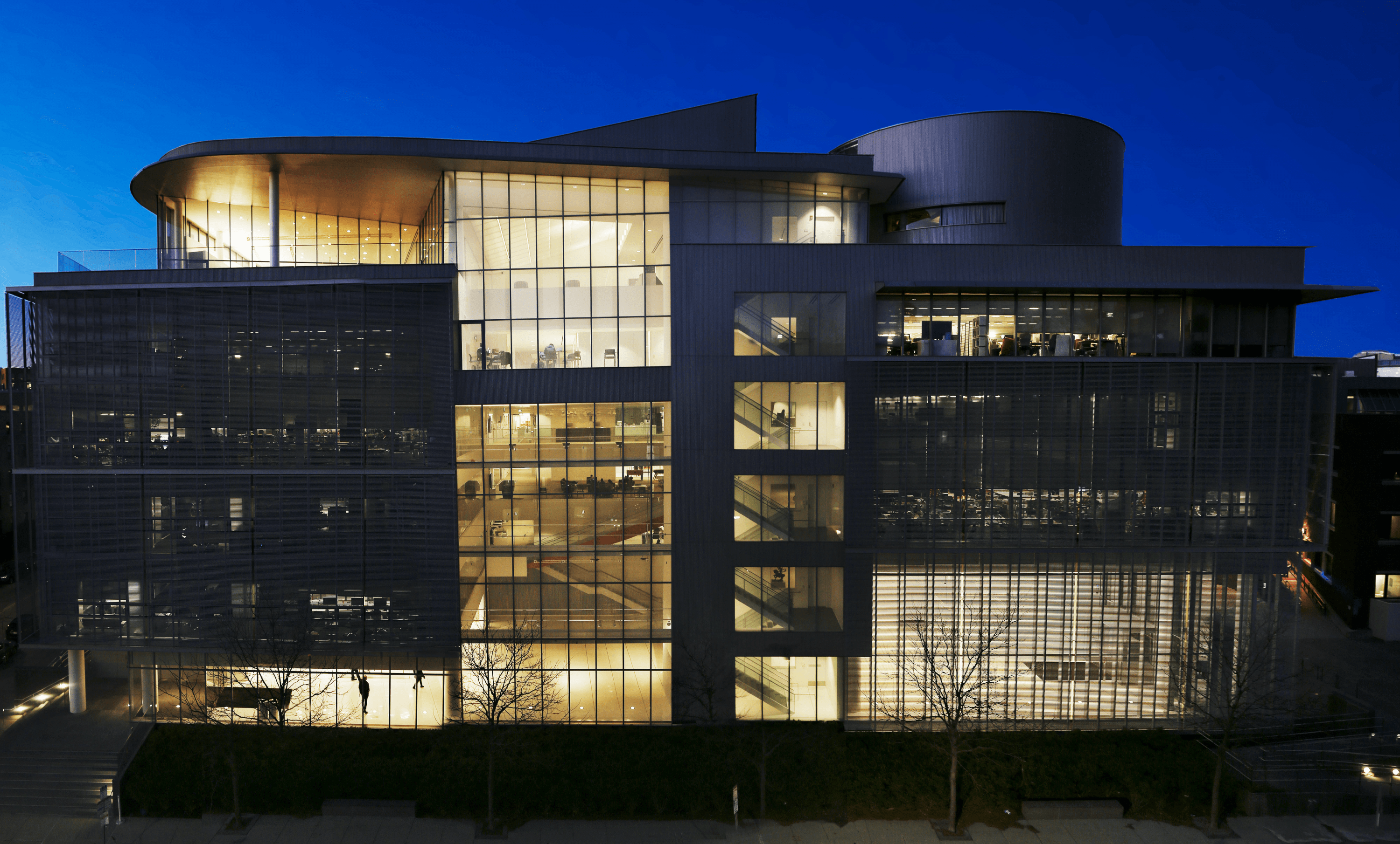 Photo of a contemporary campus building; the glass is illuminated against a night sky.