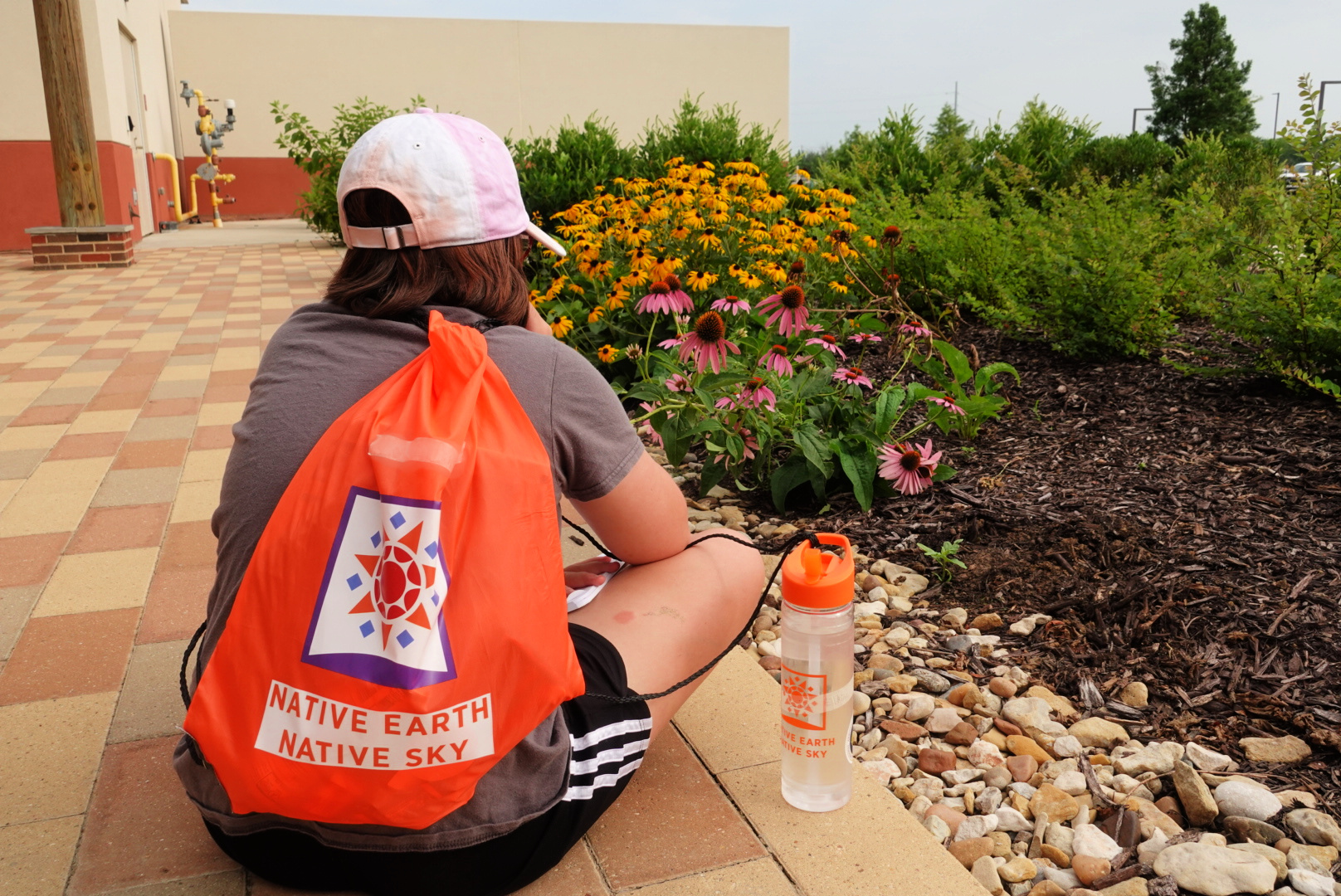 Photo of the back of a person sitting on the ground with an orange back slung over their shoulder. The bag has the Native Earth | Native Sky logo on it.