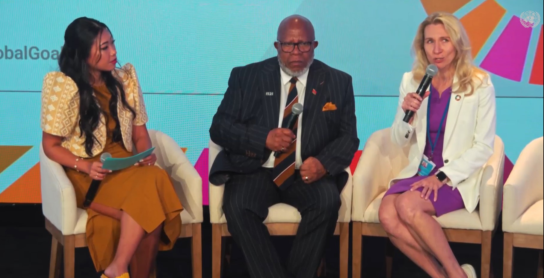 Three people with microphones sit on white chairs on stage.