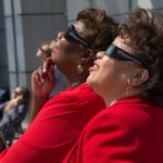 Two NASA employees wearing protective glasses to view a partial solar eclipse are smiling as they stand side-by-side and gaze up at the sky.