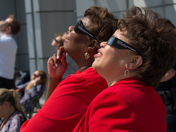 Two NASA employees wearing protective glasses to view a partial solar eclipse are smiling as they stand side-by-side and gaze up at the sky.
