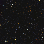 Image is filled with distant galaxies in colors of yellow-gold, red, blue, white. Lower left holds a face on spiral galaxy.