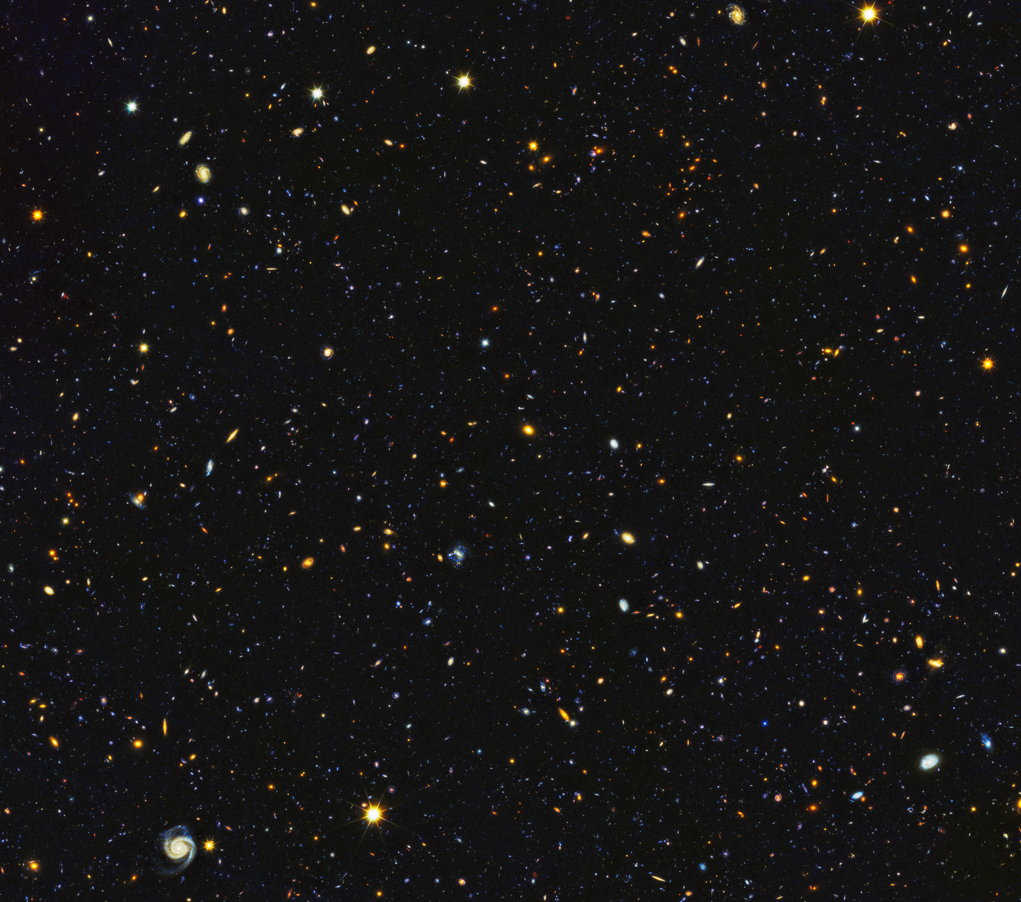 Image is filled with distant galaxies in colors of yellow-gold, red, blue, white. Lower left holds a face on spiral galaxy.