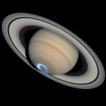 Yellow, rust, and white bands visible on Saturn. The planet is tilted, such that its rings extend from upper-left to lower-right. A blue-white, glowing ring (the auroras) is visible at the bottom of the planet's sphere. All on a black background.