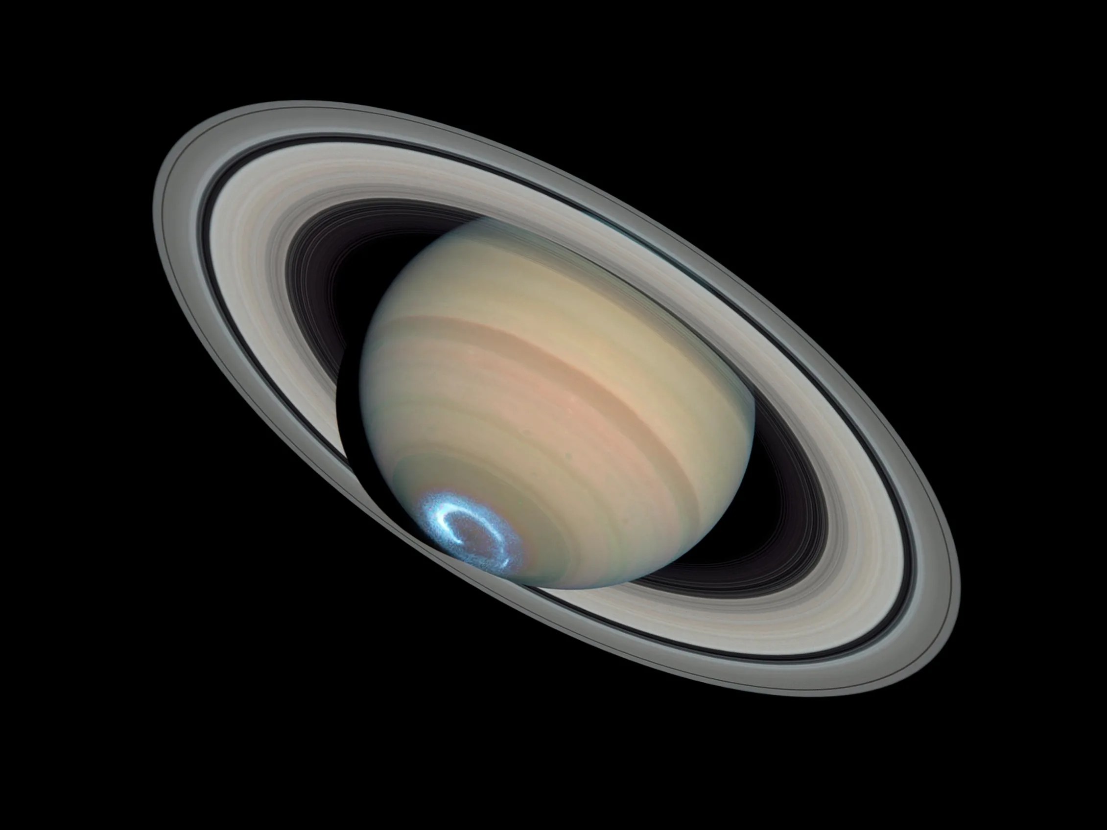 Yellow, rust, and white bands visible on Saturn. The planet is tilted, such that its rings extend from upper-left to lower-right. A blue-white, glowing ring (the auroras) is visible at the bottom of the planet's sphere. All on a black background.