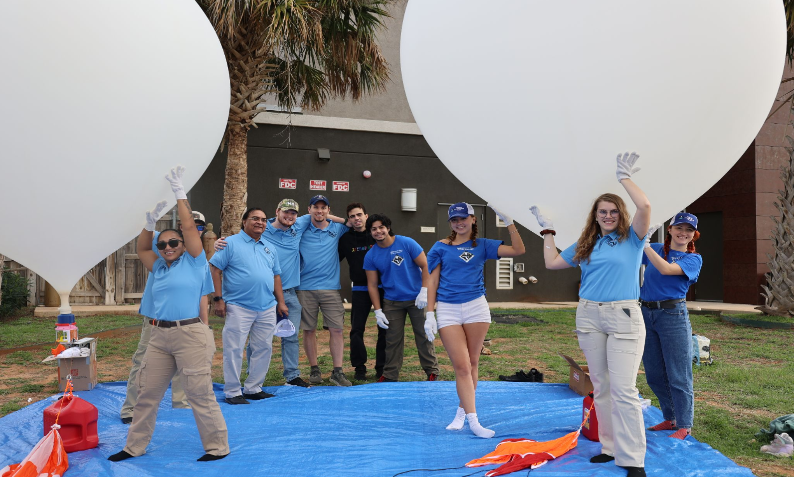 Group of people standing on a blue tarp holding large white weather balloons.