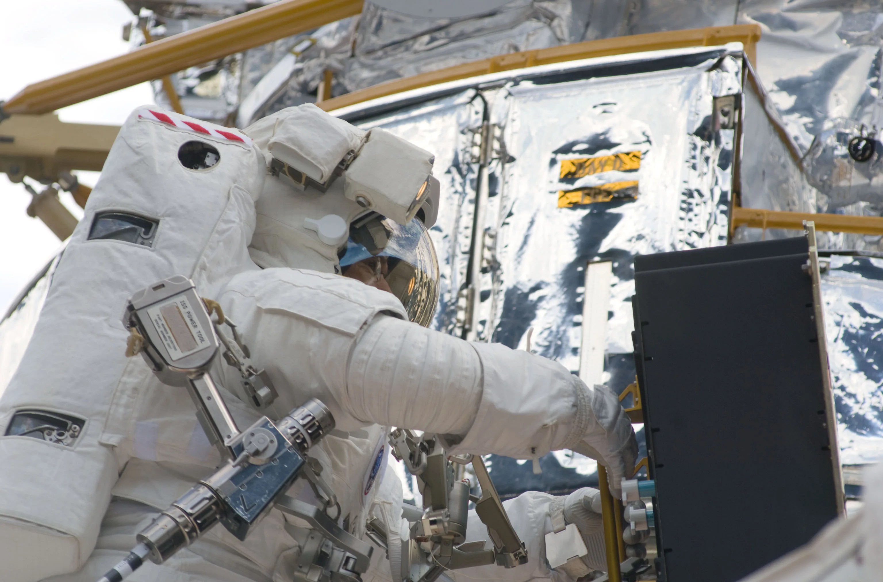 Image shows the side of an astronaut in his space suit in space. He is changing out batteries on the side of the Hubble Space Telescope. The picture is bright, both astronaut and Hubble were in the sun when this was taken. The astronaut's pistol grip tool, which looks like an electric screwdriver, rests at his side.