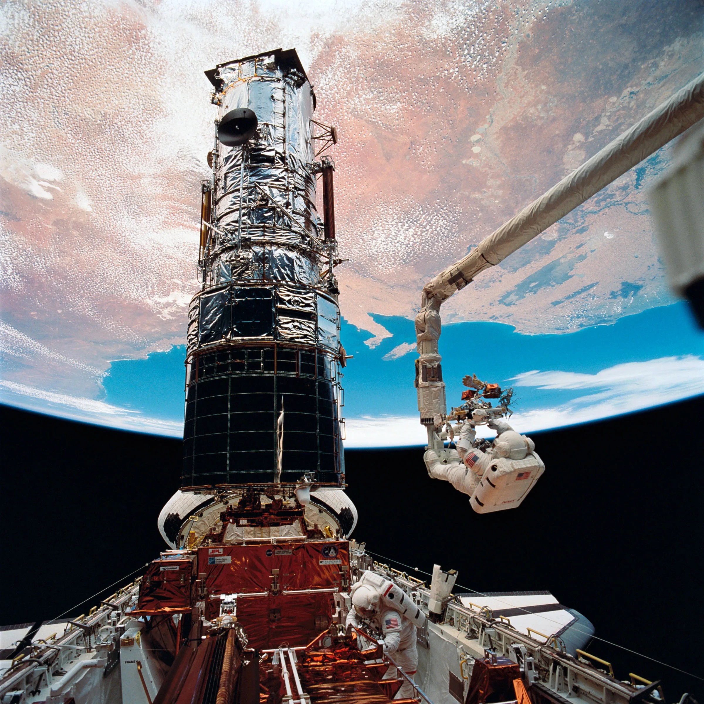 Two astronauts service Hubble, one standing in the space shuttle cargo bay and one attached to a robotic arm, with the Earth in the background in the upper half of the picture.