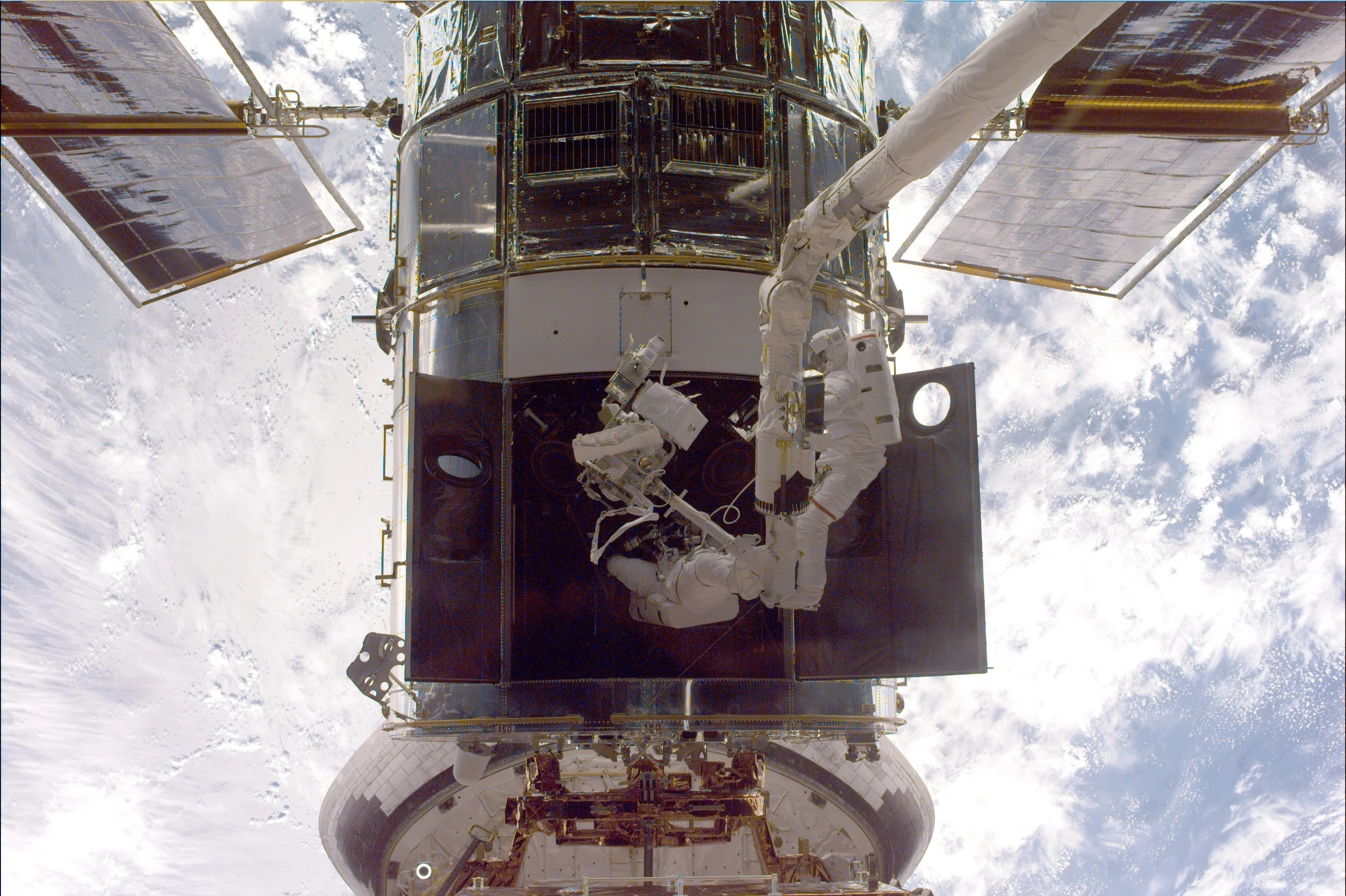 Two astronauts, attached to a robotic arm, service Hubble above the Earth, which takes up the whole background.