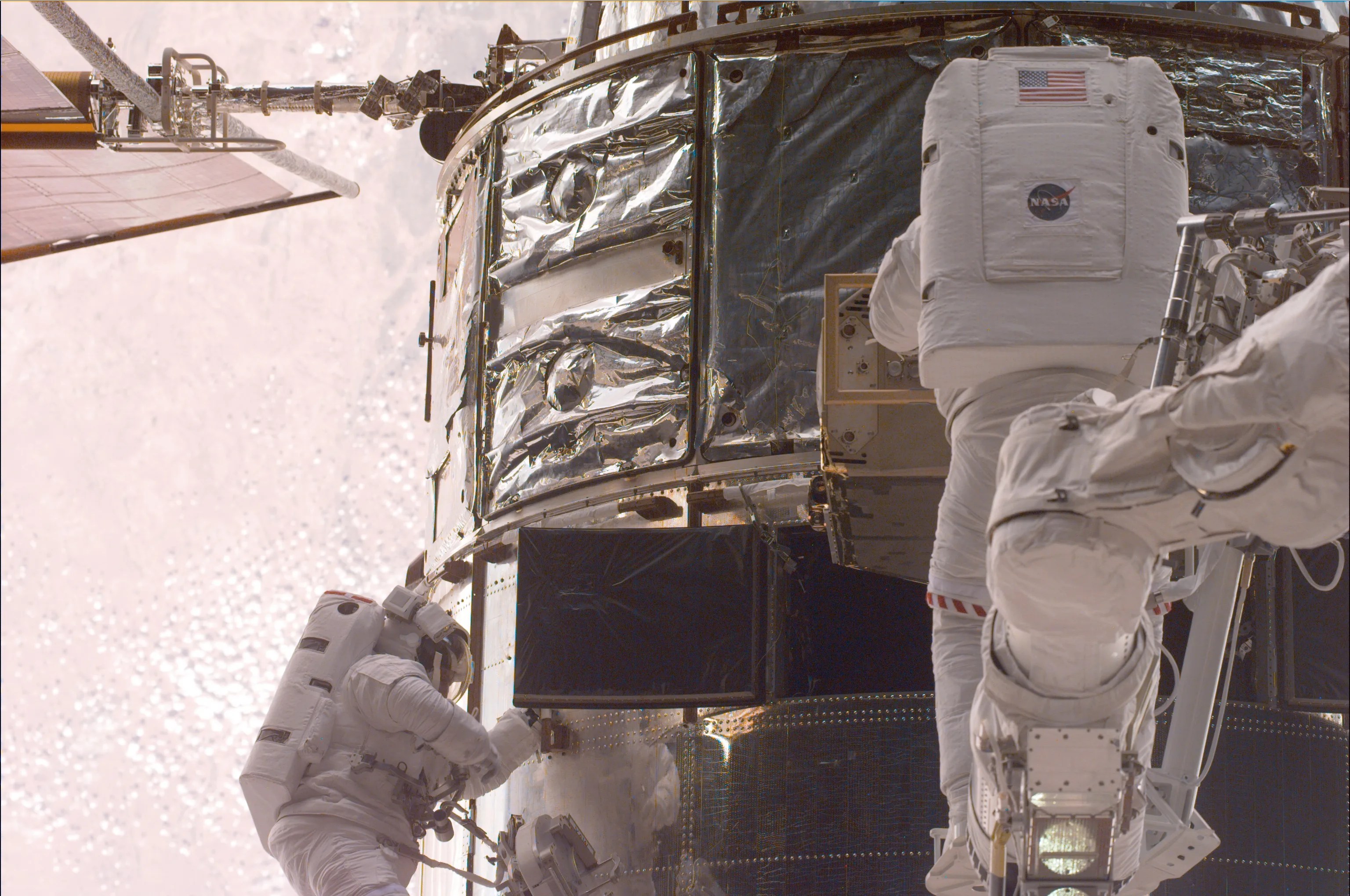 A close-up view of two astronauts servicing Hubble, one tethered to the telescope on the left and one attached to a robotic arm in the center. The ground below on Earth takes up the entire background.