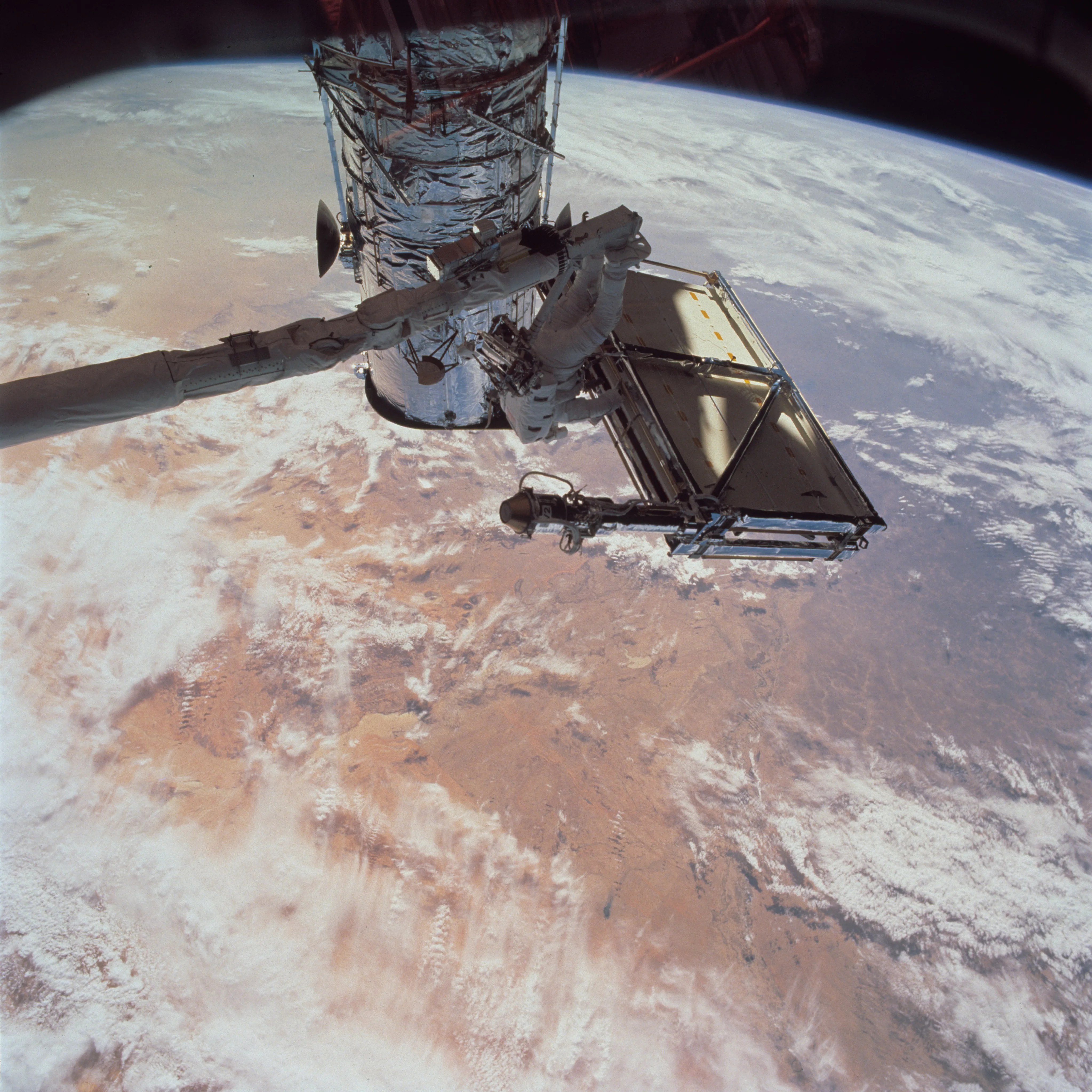 An astronaut attached to a robotic arm prepares to re-open Hubble's folded-up solar panel "wings. The Earth below takes up most of the background.