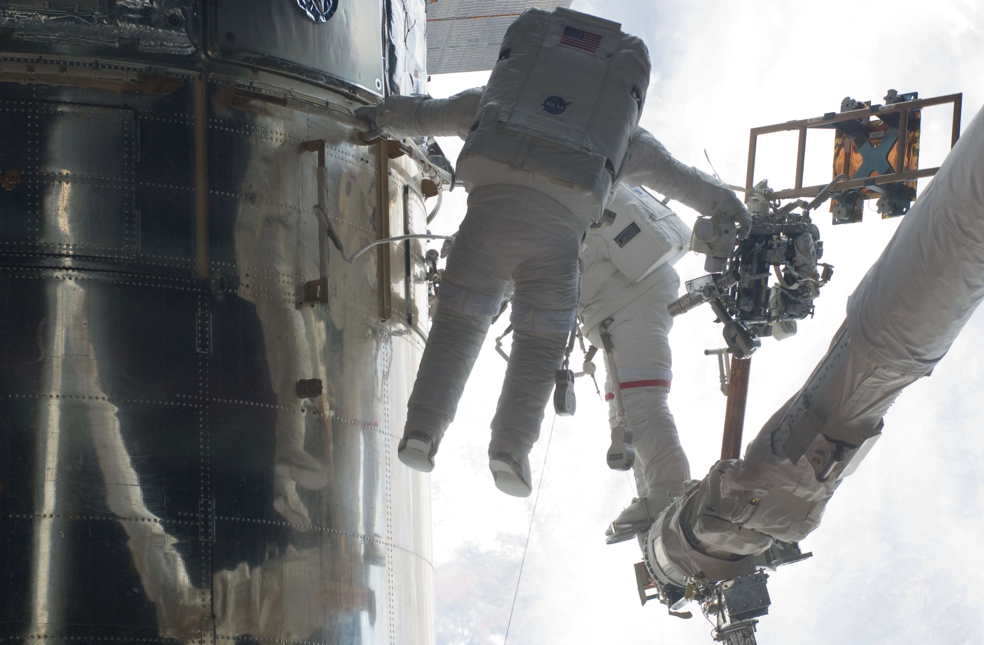 Two astronauts servicing Hubble using a robotic arm. Clouds above Earth's surface behind the telescope create the background.