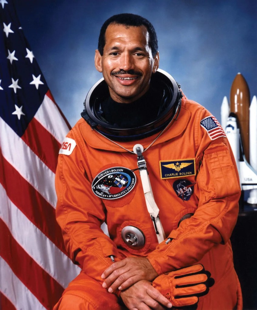 Photo of astronaut Charles Bolden in his orange spacesuit. American flag is behind him.