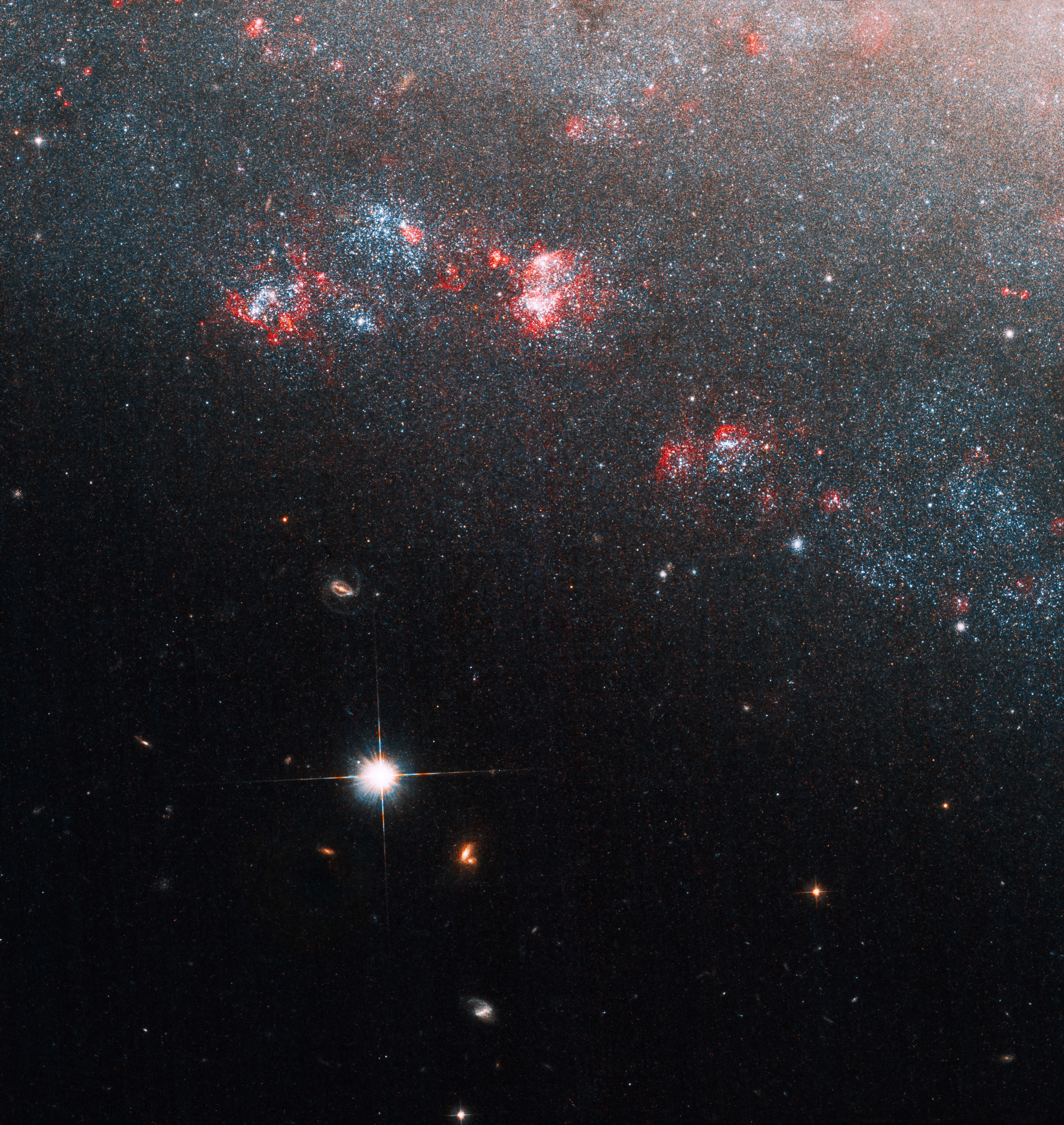 A haze of stars fills the top and upper right corner of NGC 247/Caldwell 62. The area is dotted with bright pinkish-red and blue gas clouds. The lower-left half of the image is black and dotted with one very bright star and a few dimmer ones.