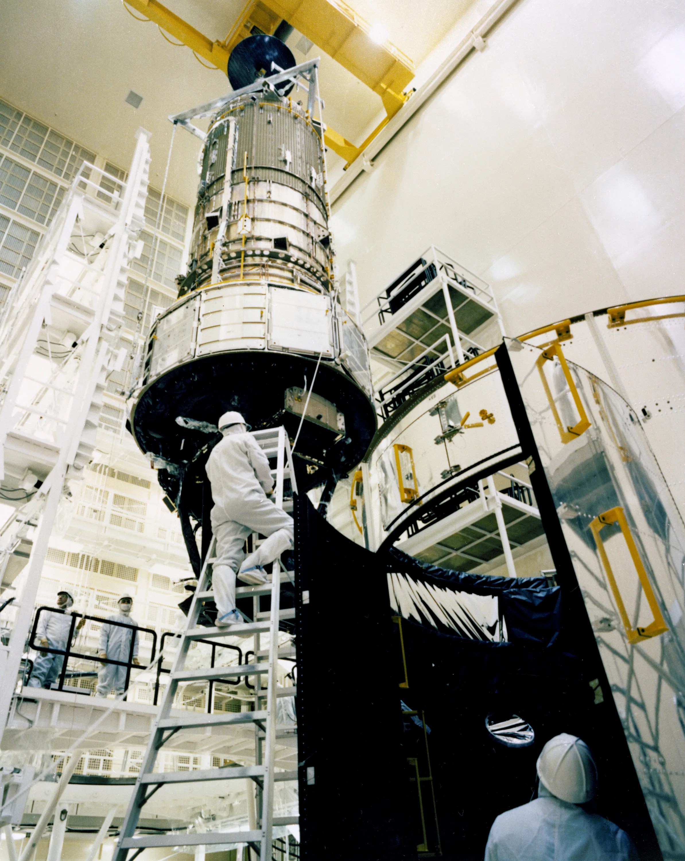 A white-garbed worker wearing a helmet climbs a ladder to reach a portion of the Hubble Space Telescope under assembly in a clean room.