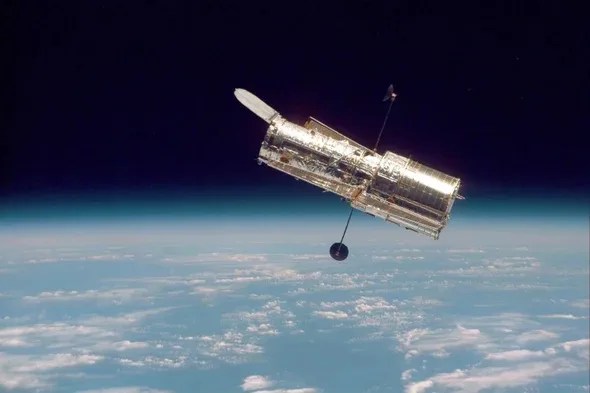 Image of Hubble seen floating above the Earth in orbit. Under Hubble, the blue colored landmass is visible through the clouds. On the upper half of the image, the darkness of space is seen.