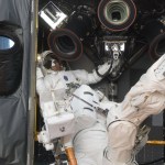 Astronaut Mike Massimino inside one of Hubble's instrument bays (left of center). Instrument Specialists, Michael Good (center) is standing on the Manipulator Foot Restraint, which is connected to the Shuttle Remote Manipulator System/Canadarm Latching End Effector. The Canadarm is white and extends from the upper right corner to the bottom center.