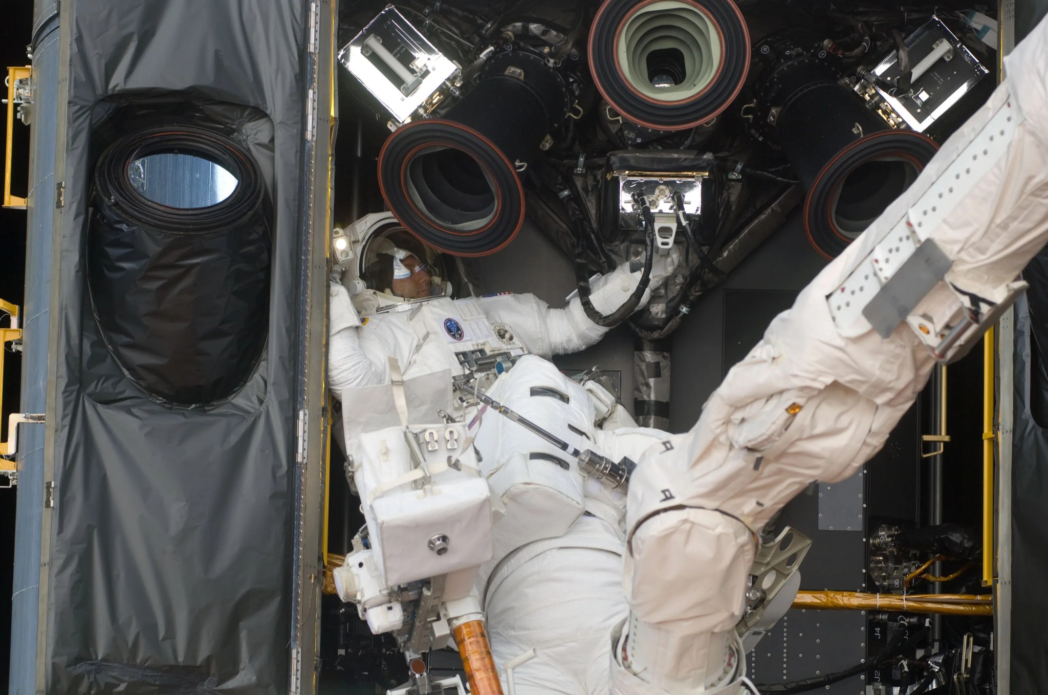 Astronaut Mike Massimino inside one of Hubble's instrument bays (left of center). Instrument Specialists, Michael Good (center) is standing on the Manipulator Foot Restraint, which is connected to the Shuttle Remote Manipulator System/Canadarm Latching End Effector. The Canadarm is white and extends from the upper right corner to the bottom center.