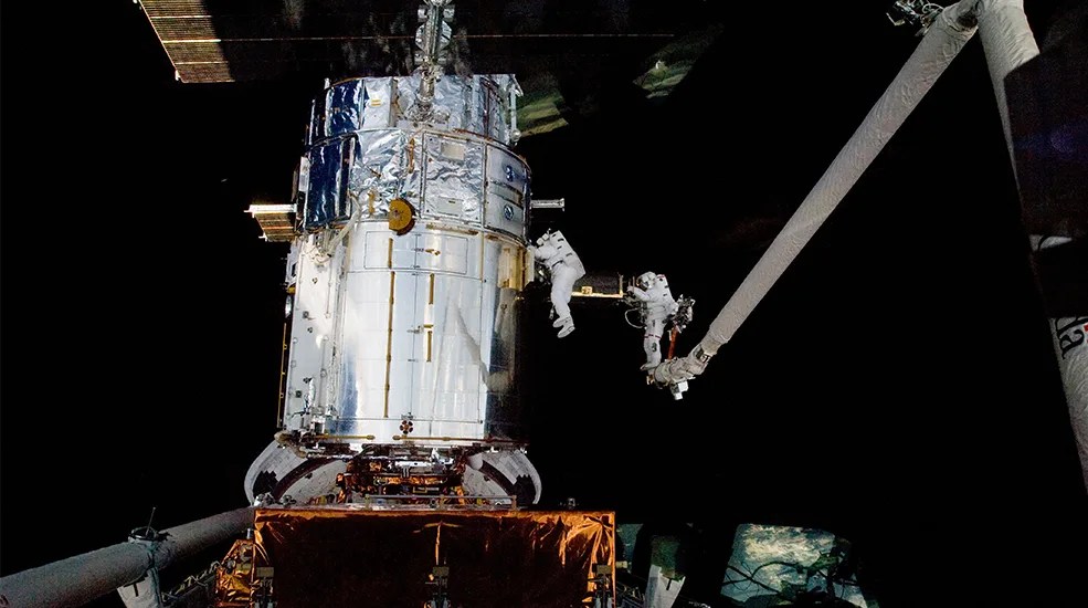 2 astronauts in their spacesuits are seen floating near the side of Hubble. Hubble, bright silver, from its reflection of the sun is many times larger than the two astronauts. On the right of the image the CANADARM is seen.