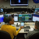 A photograph of a man and woman sitting in Hubble's "Space Telescope Operations Control Center" also called the STOCC. They are sitting next to each other, with many computer screens with data and information from the spacecraft.