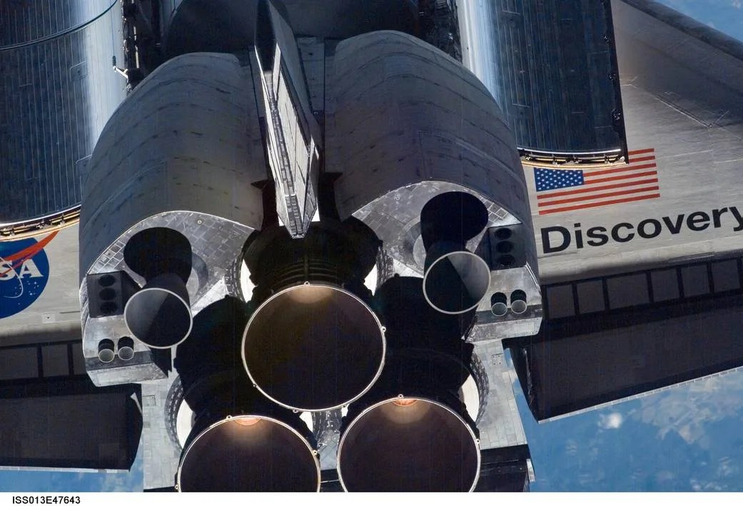 Closeup image of the Space Shuttle Discovery. In this image you can only see its powerful black engines, while on the right wing the American Flag is present along with the words "Discovery" under it. The left wing is mostly cut off from the image, but you can see half of the blue NASA Meatball. Behind the spacecraft you can just make out the blue Earth below.