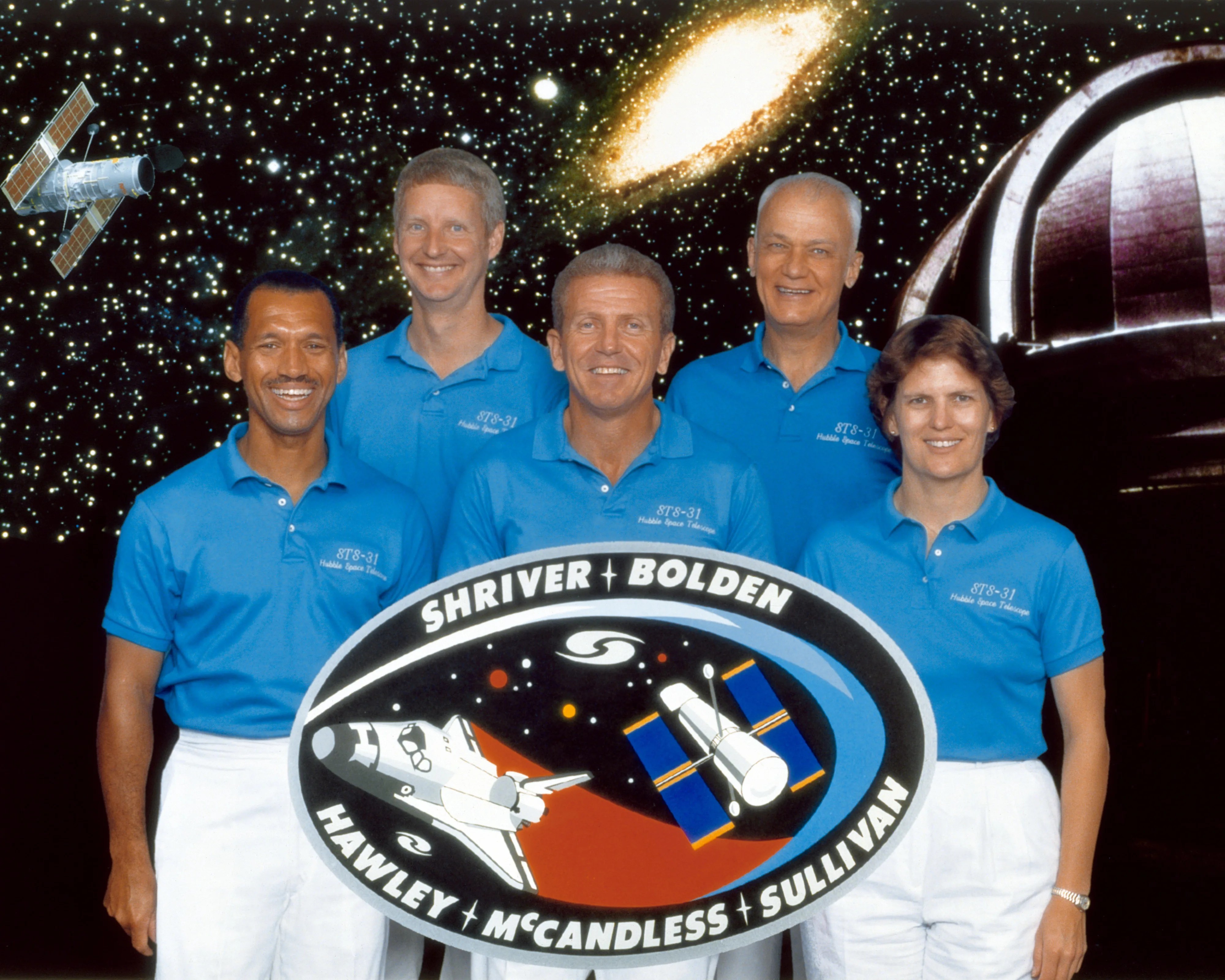 Five astronauts--four men and one woman--stand in a line in front of a starry backdrop, all wearing light blue T-shirts and white pants.