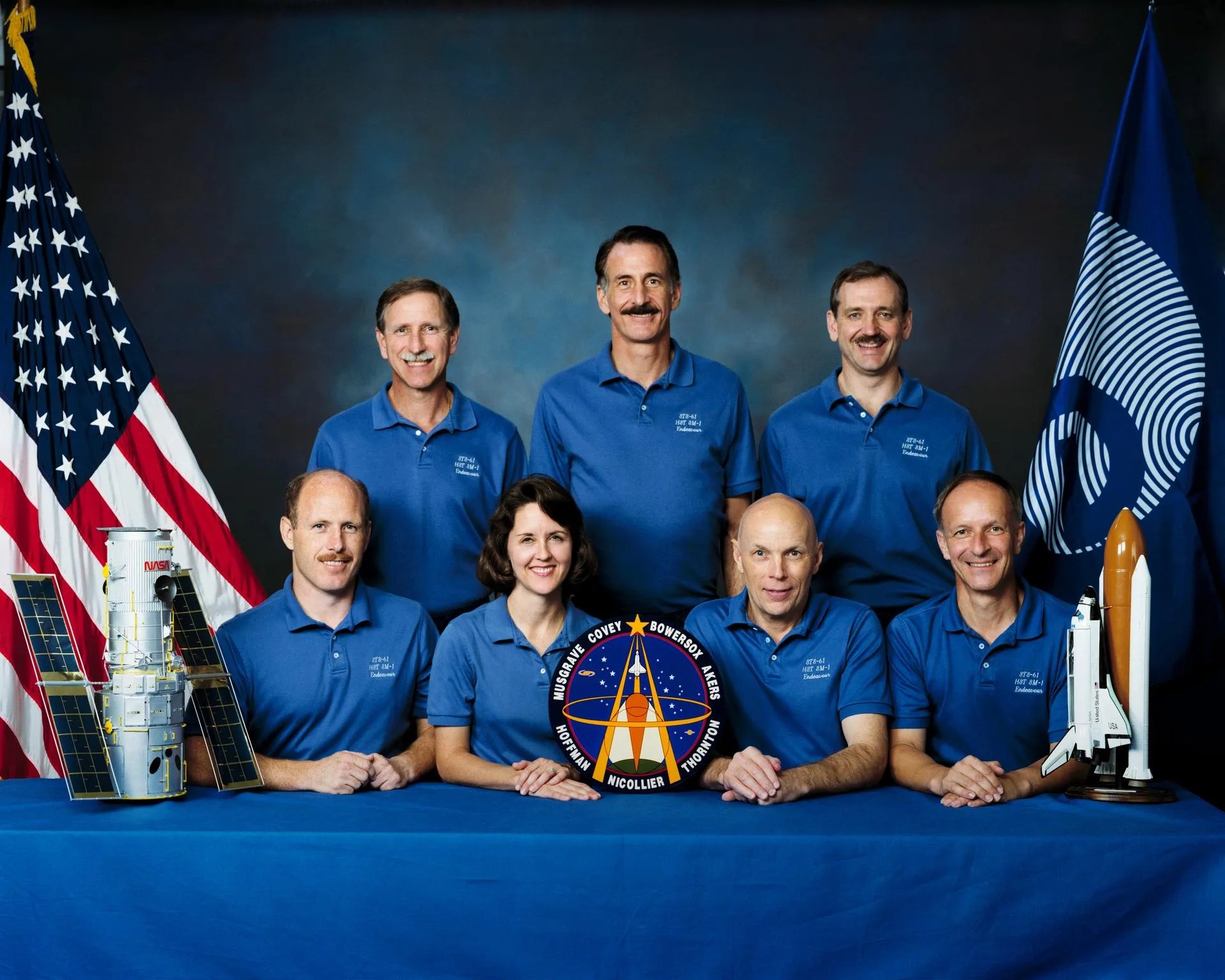 Seven astronauts wear blue T-shirts: three men standing in the back row, and three men and one woman sitting at the table in the front row. On either side of them are US and ESA flags, and models of the space shuttle and the Hubble telescope.