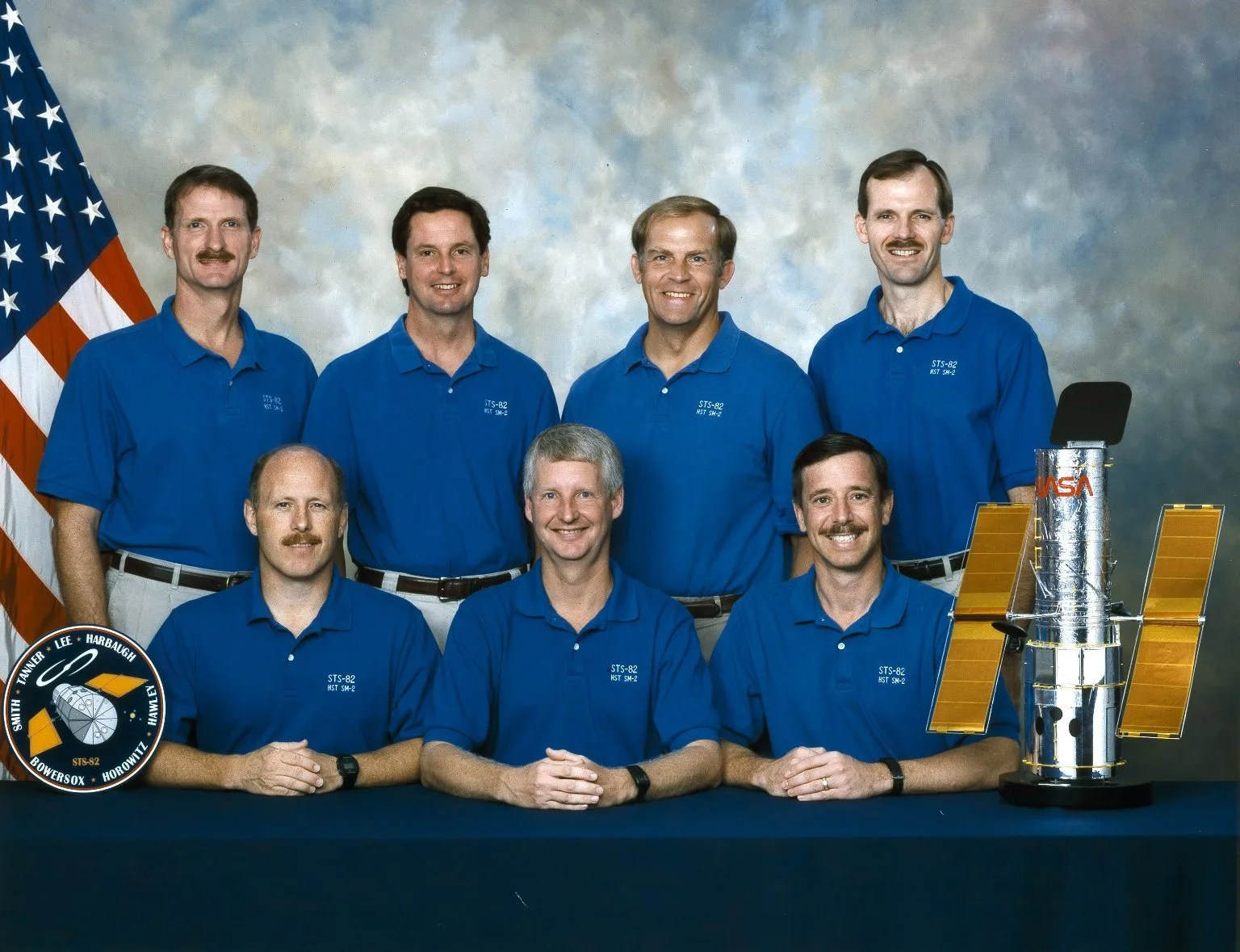 Seven astronauts wear blue T-shirts and white pants; four men stand in the back row, and three sit at at able in the front row. On the far left is a US flag, and on the right is a model of Hubble.