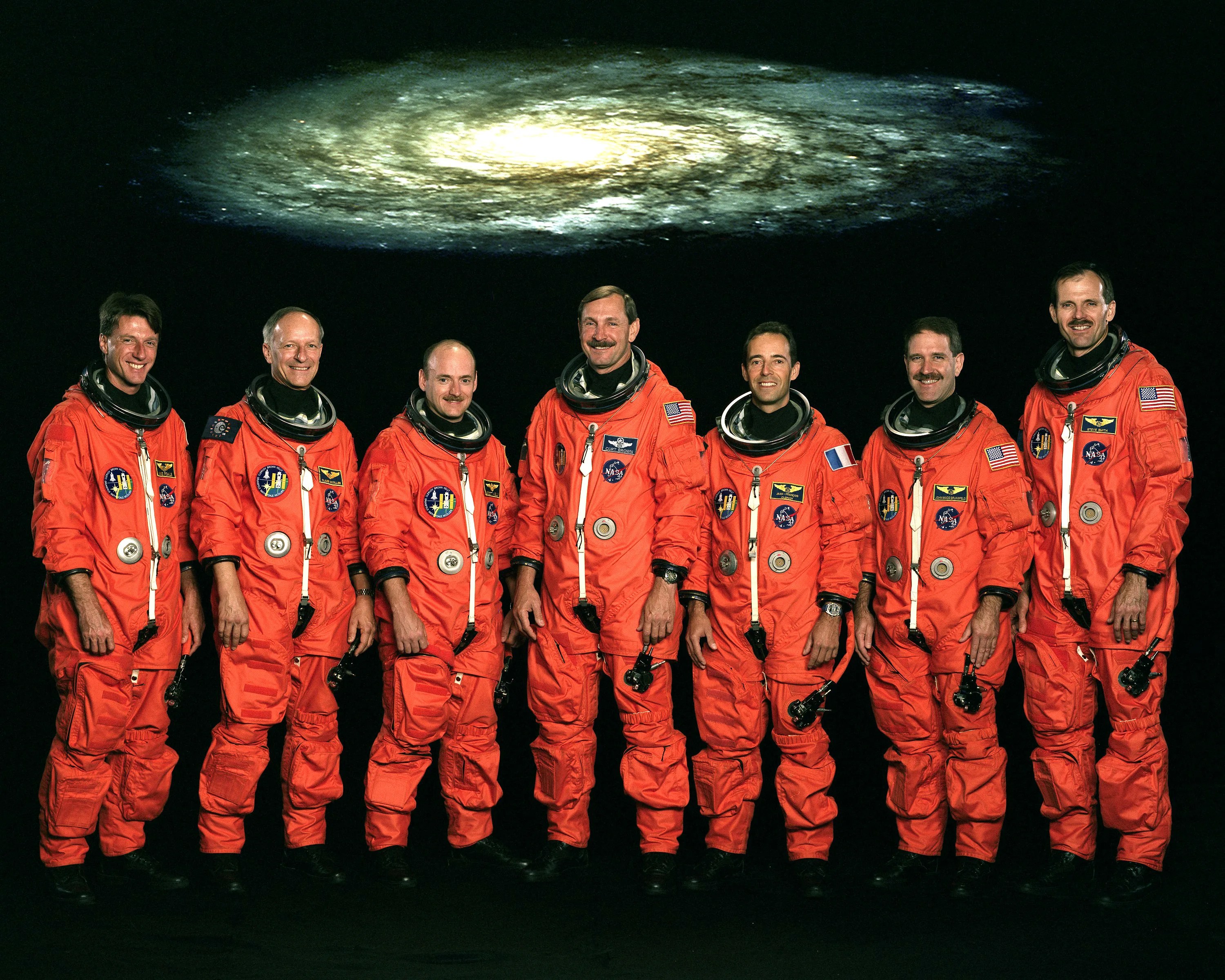 Seven astronauts in orange space suits stand in a line. A spiral galaxy hovers above their heads in the backdrop.