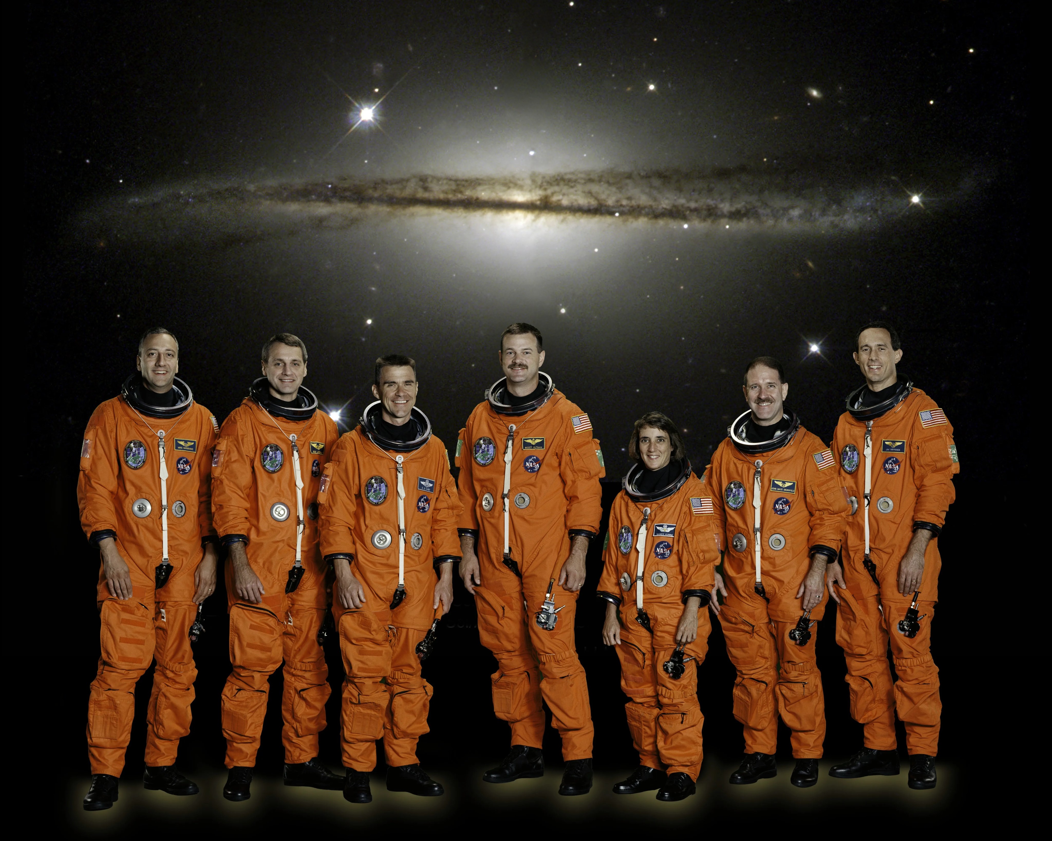 Seven astronauts in orange space suits stand in a line. The dusty arm of a spiral galaxy disc hovers above their heads in the backdrop.