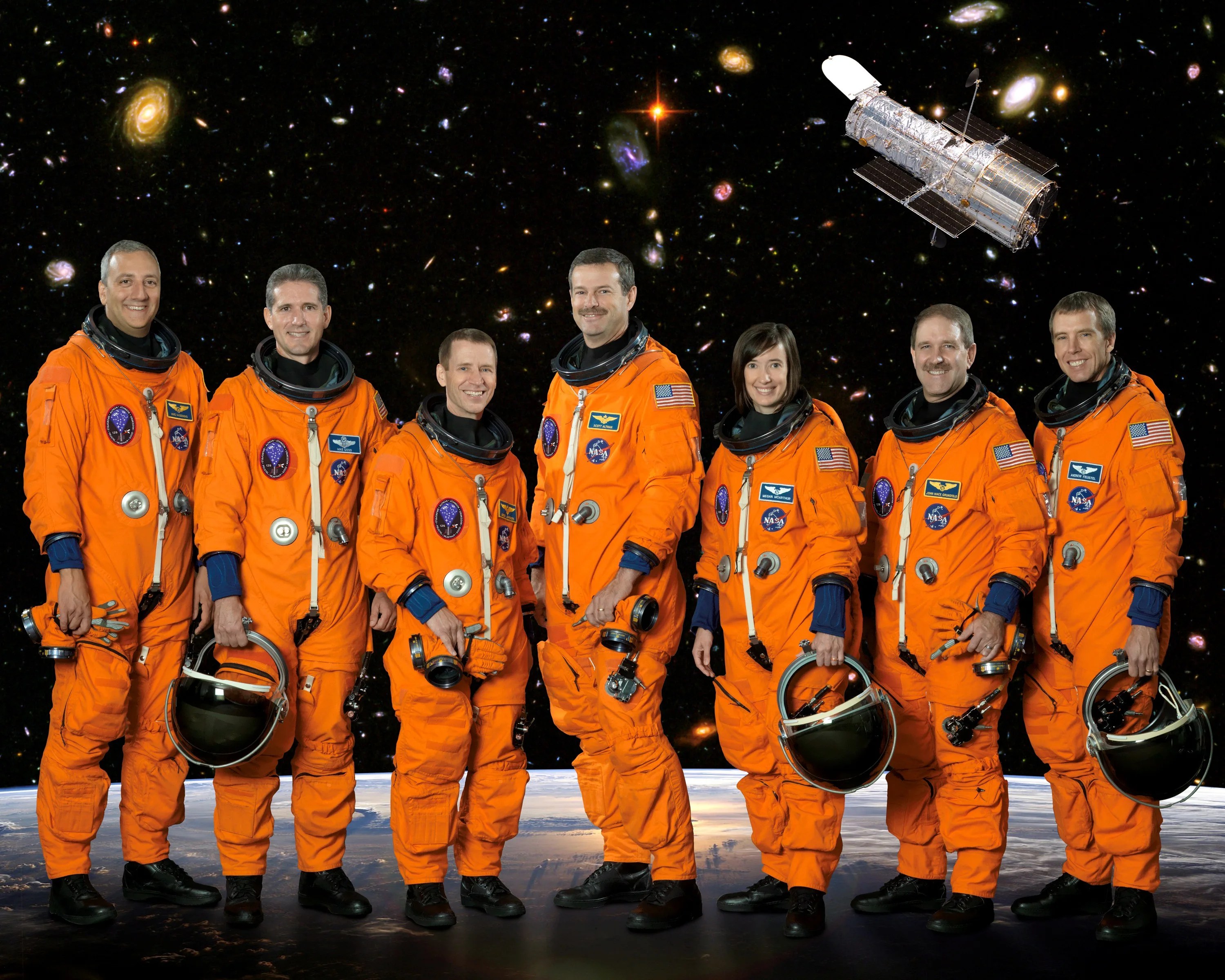 Seven astronauts--six men and one woman--stand in a line in orange astronaut suits, a few of them holding their helmets, with an image of the Hubble Ultra Deep Field in the background.