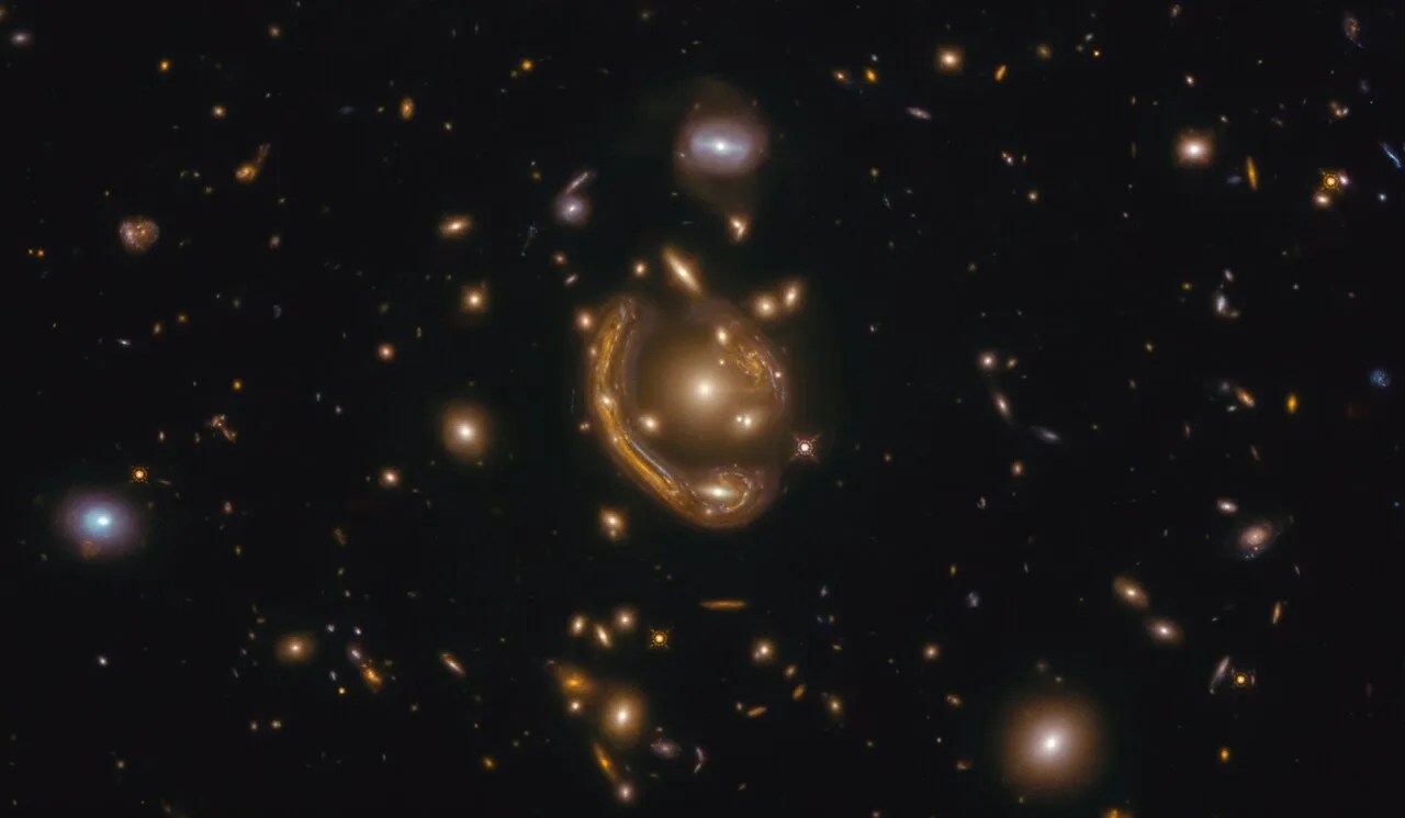 Hubble image of an "Einstein Ring." In this image we see dozens and dozens of bright points of light, all of which are galaxies. In the middle however, we see a stretched out ring of light which almost makes an entire circle, or ring. In the middle of the ring there are more points of light clustered together.