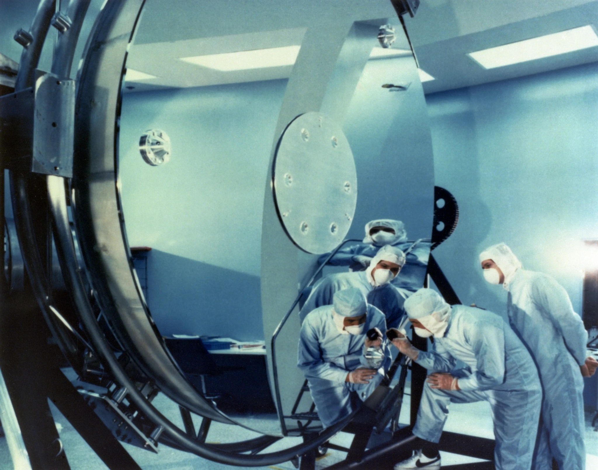 Engineers in full-body bunny suits closely inspect the giant mirror of the Hubble Space Telescope.