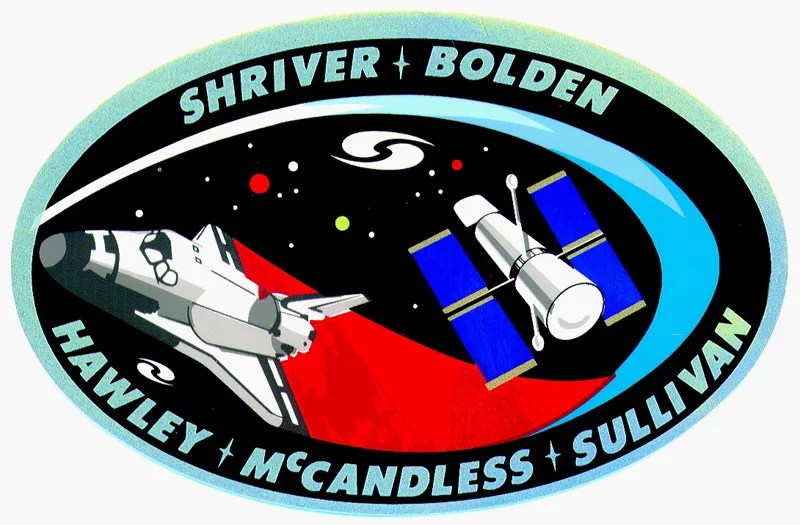 Hubble Crew patch. Oval patch with "Shriver, Bolden, Hawley, McCandless, Sullivan" written on the border. These are the names of the astronauts on the mission. Inside the patch we see the Shuttle swooping in front of a black background, with Hubble in the image as well.