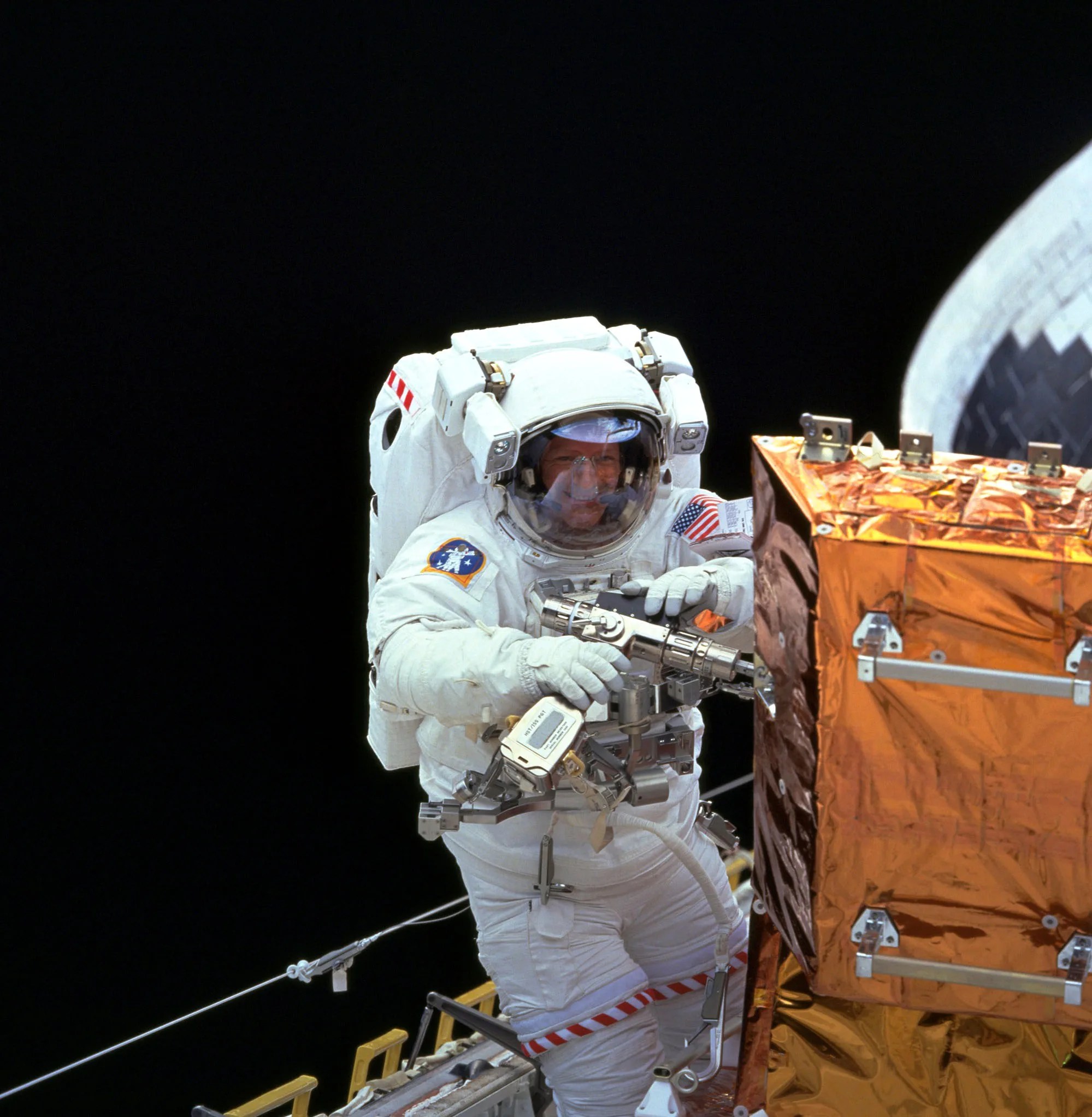 An astronaut holding the Pistol Grip Tool, which looks like an electric screwdriver. The astronaut's face is visible through his visor, he is smiling. He stands next to a large gold box with a bit of the Shuttle visible behind him.