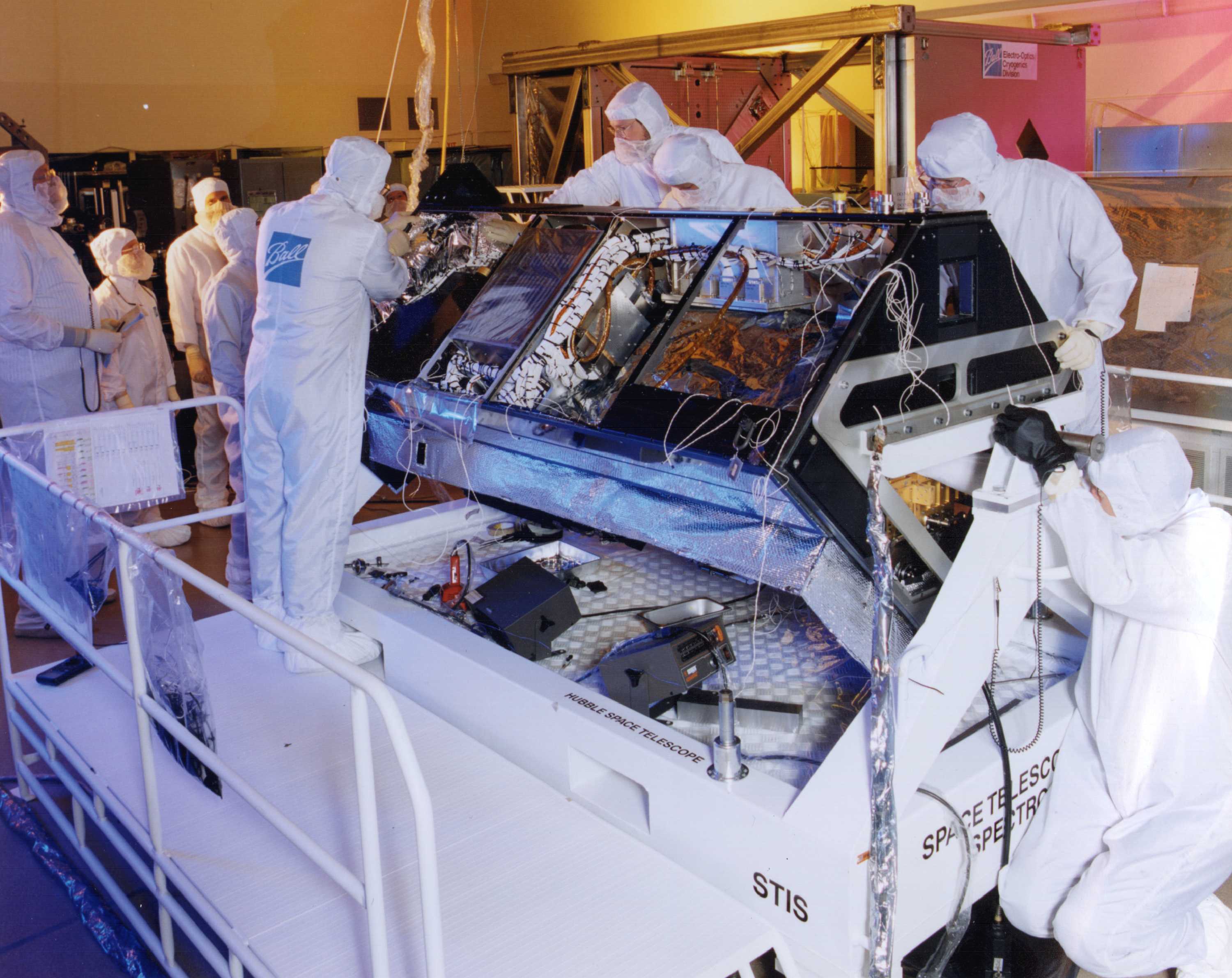 Workers in cleanroom suits work on a large box with many wires sticking out that is the Space Telescope Imaging Spectrograph
