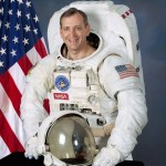 Photo of astronaut Thomas Akers in his spacesuit. American flag is behind him.