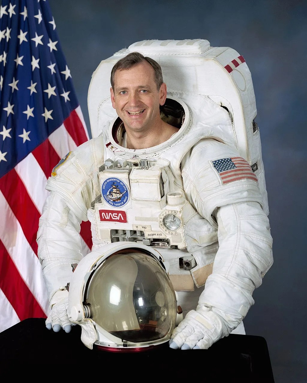 Photo of astronaut Thomas Akers in his spacesuit. American flag is behind him.