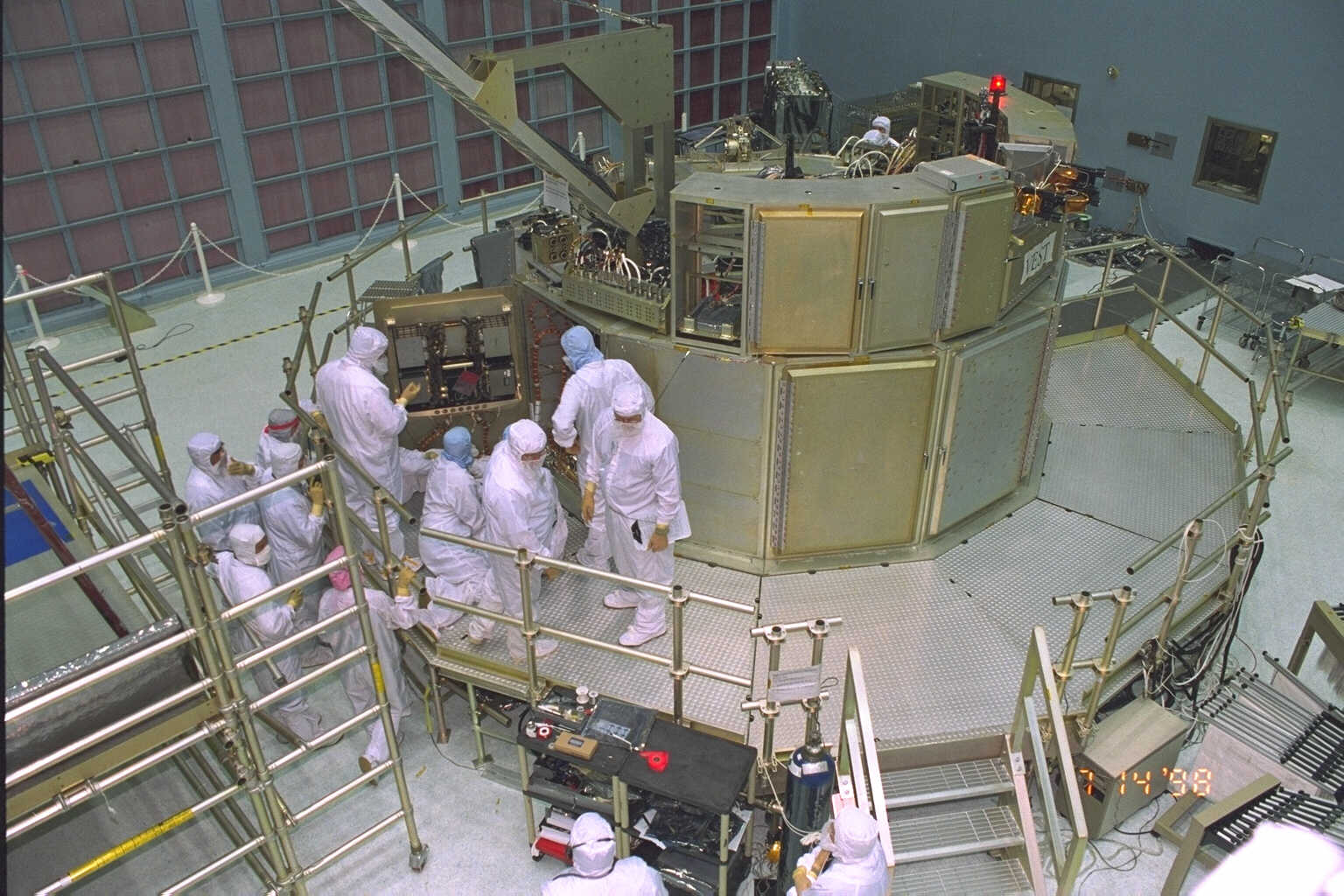 Astronauts standing around the VEST, in their full-body bunny suits. astronauts are seen receiving instruction from Hubble project personnel on various spacecraft subsystems.