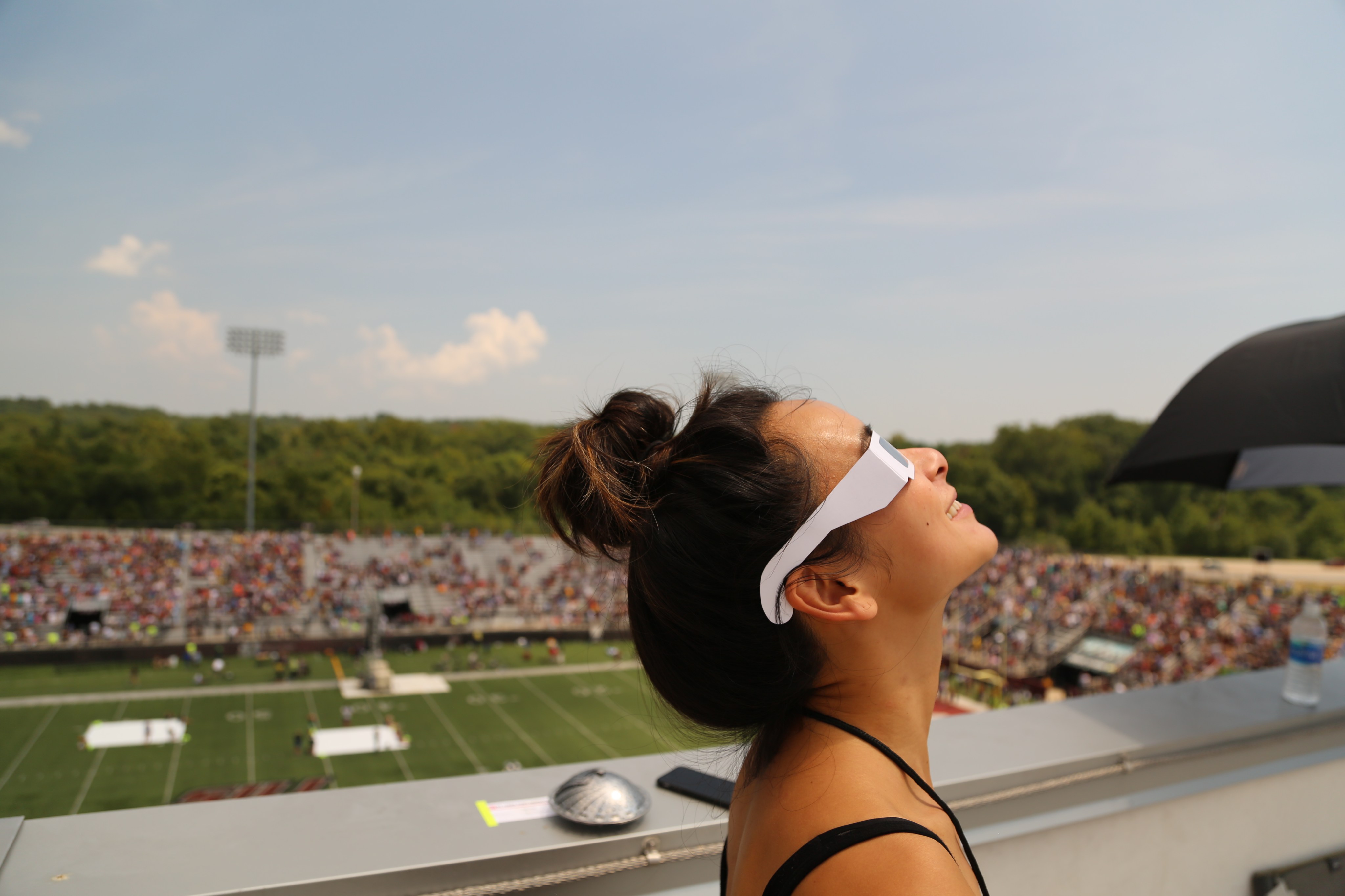 A woman, standing in profile, wears white eclipse glasses. She's looking up toward the sky. Behind her, out of focus, is stands mostly filled with people and a football field.