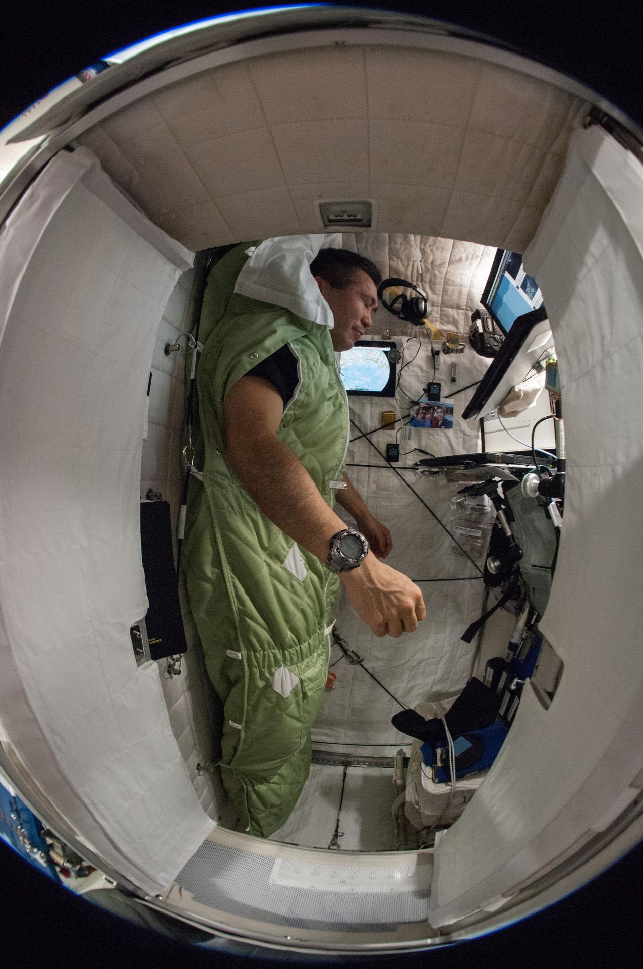 A man is inside the International Space Station. He appears to be upright, but is wearing a padded green sleeping bag that covers his entire body except his arms, which poke out through holes in the side. His eyes are closed.